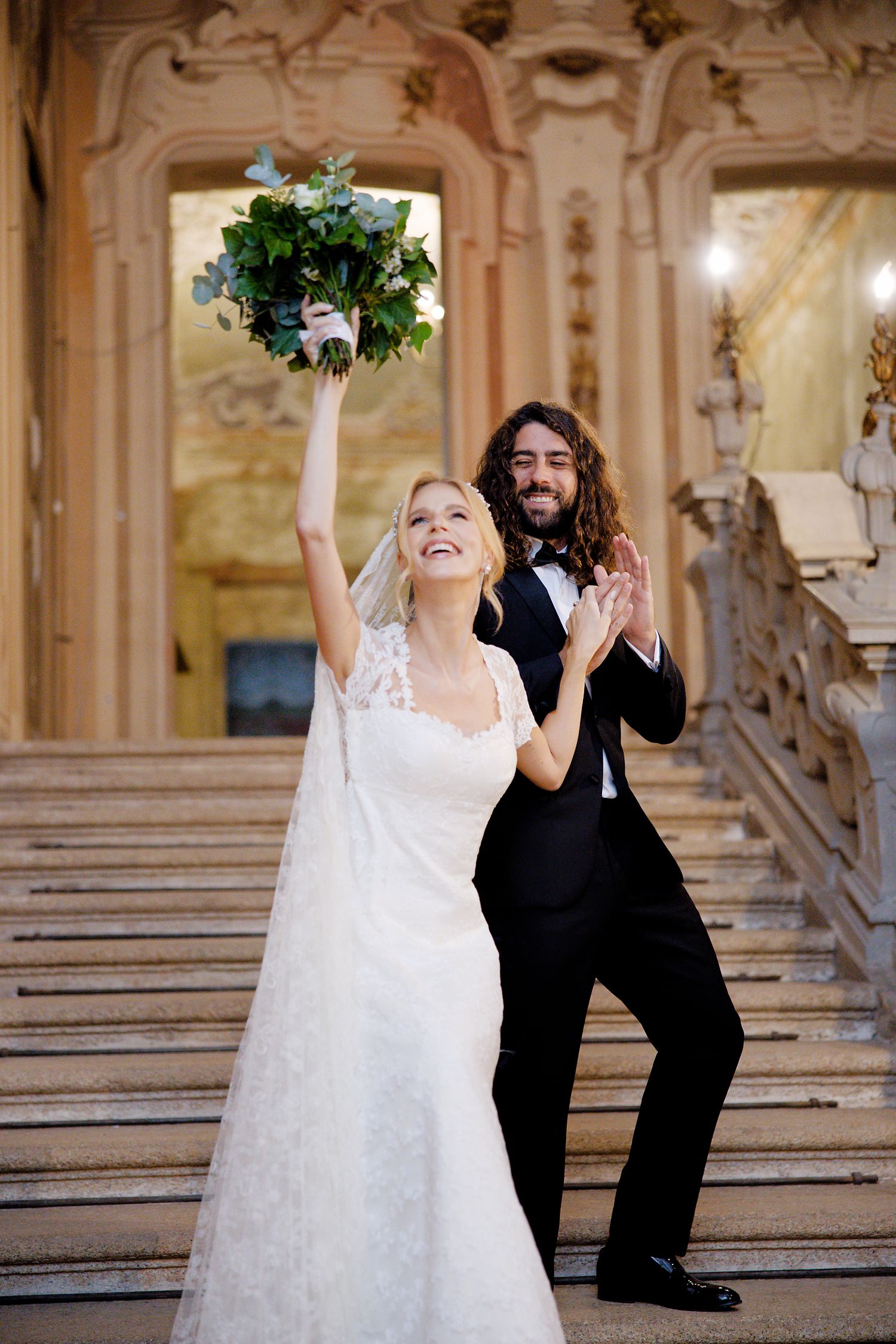 A joyful wedding couple celebrating on an elegant marble staircase with a held-up floral bouquet.
