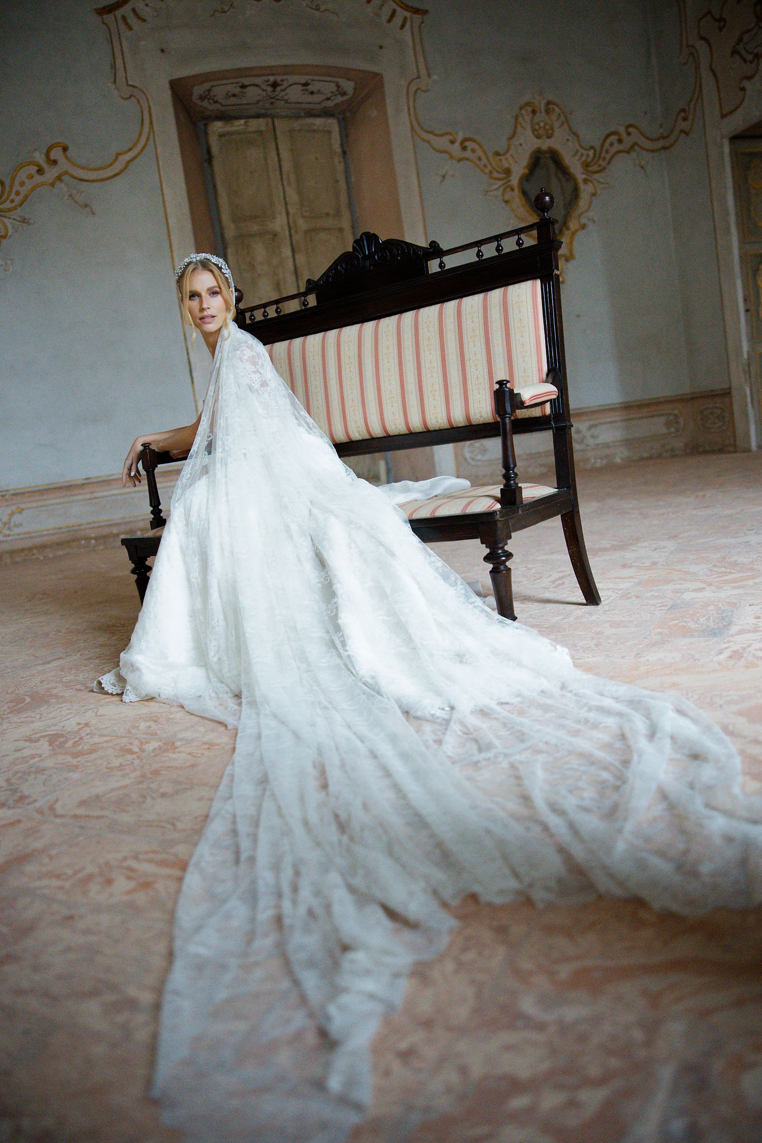 A bride in an elegant white lace wedding gown with a dramatic long train poses in a grand ornate room.