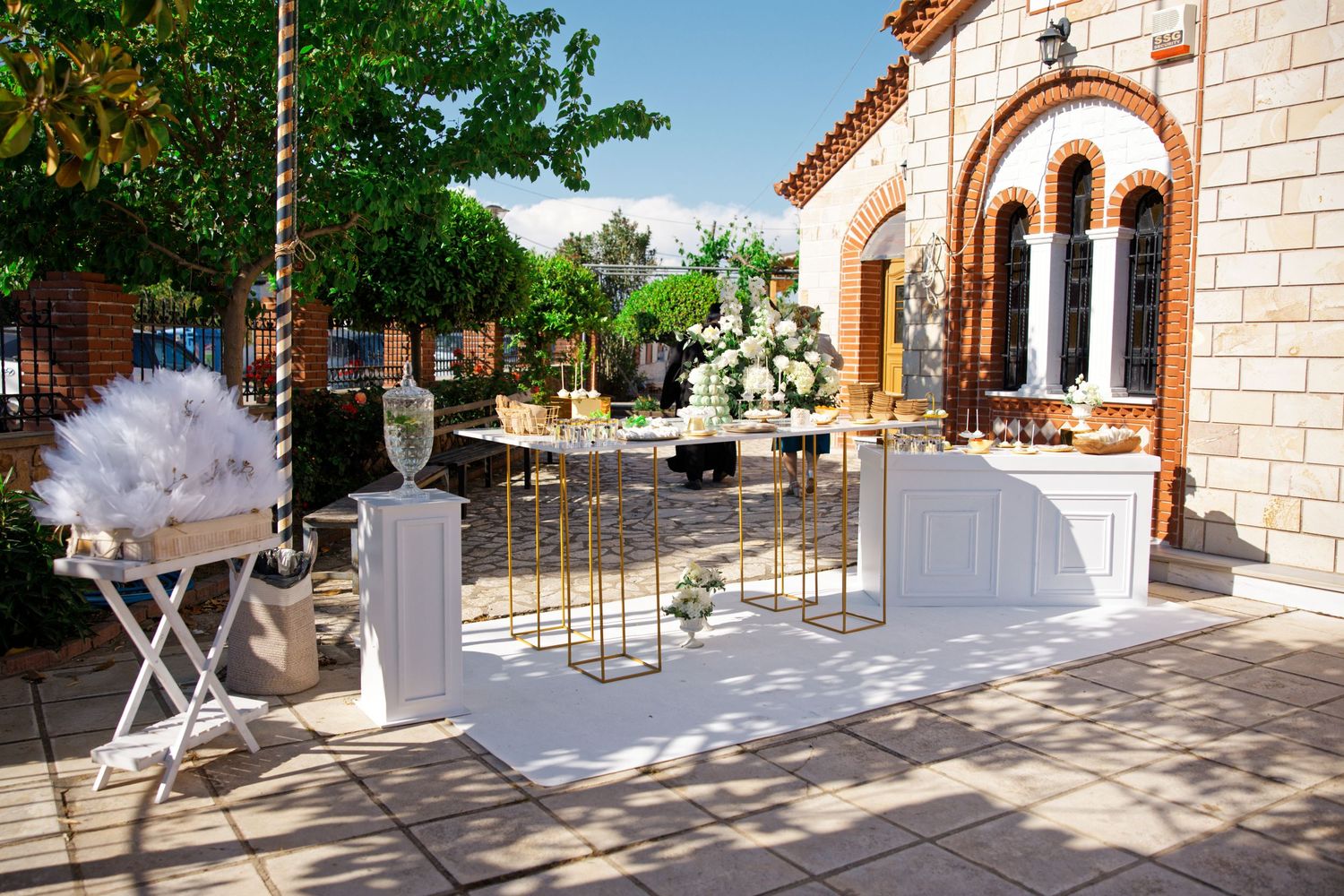 A white wedding ceremony setup with decorative elements on a sunny outdoor patio near a stone building.