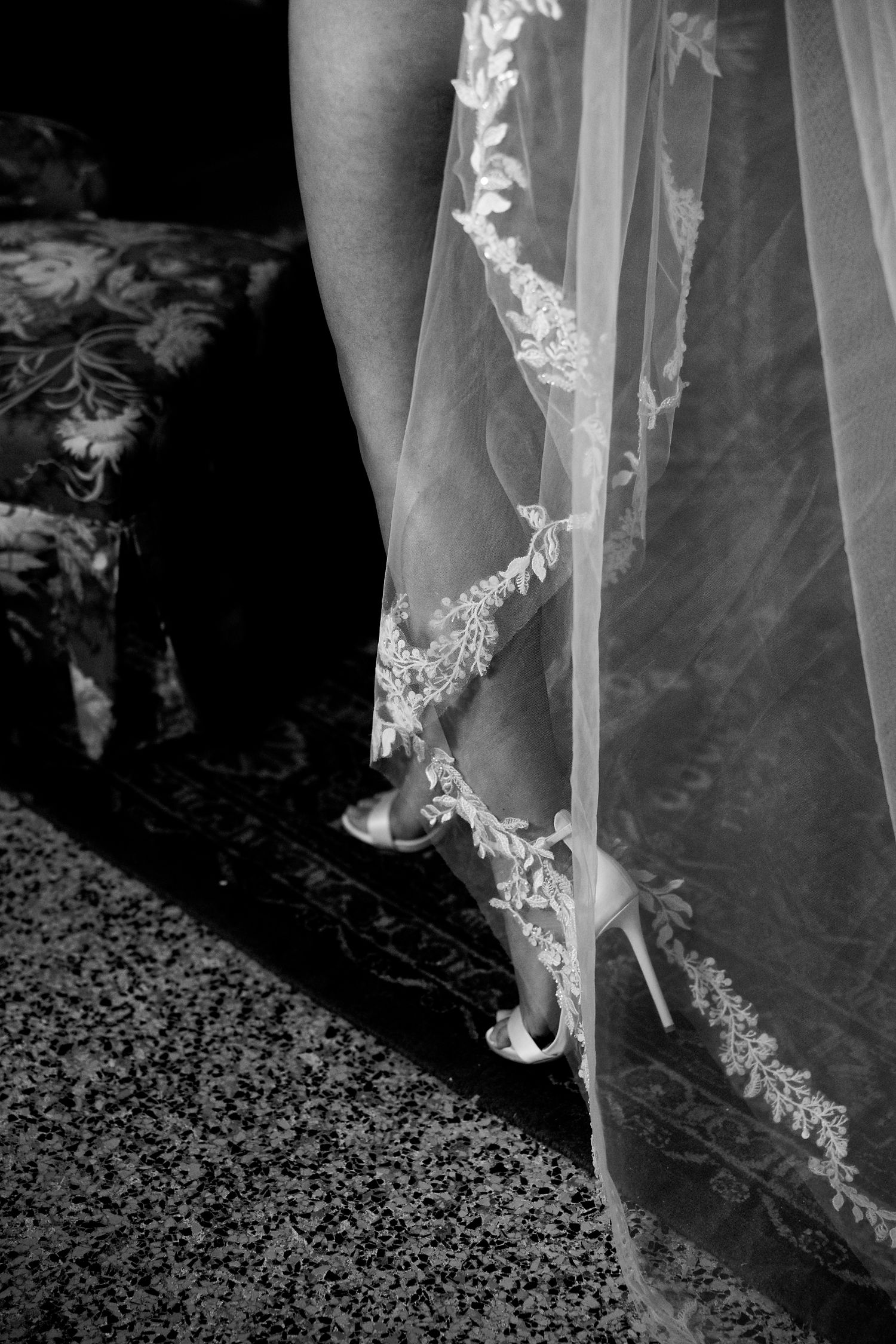 Close-up black and white photo of delicate lace wedding veil detail and shoes on patterned carpet.