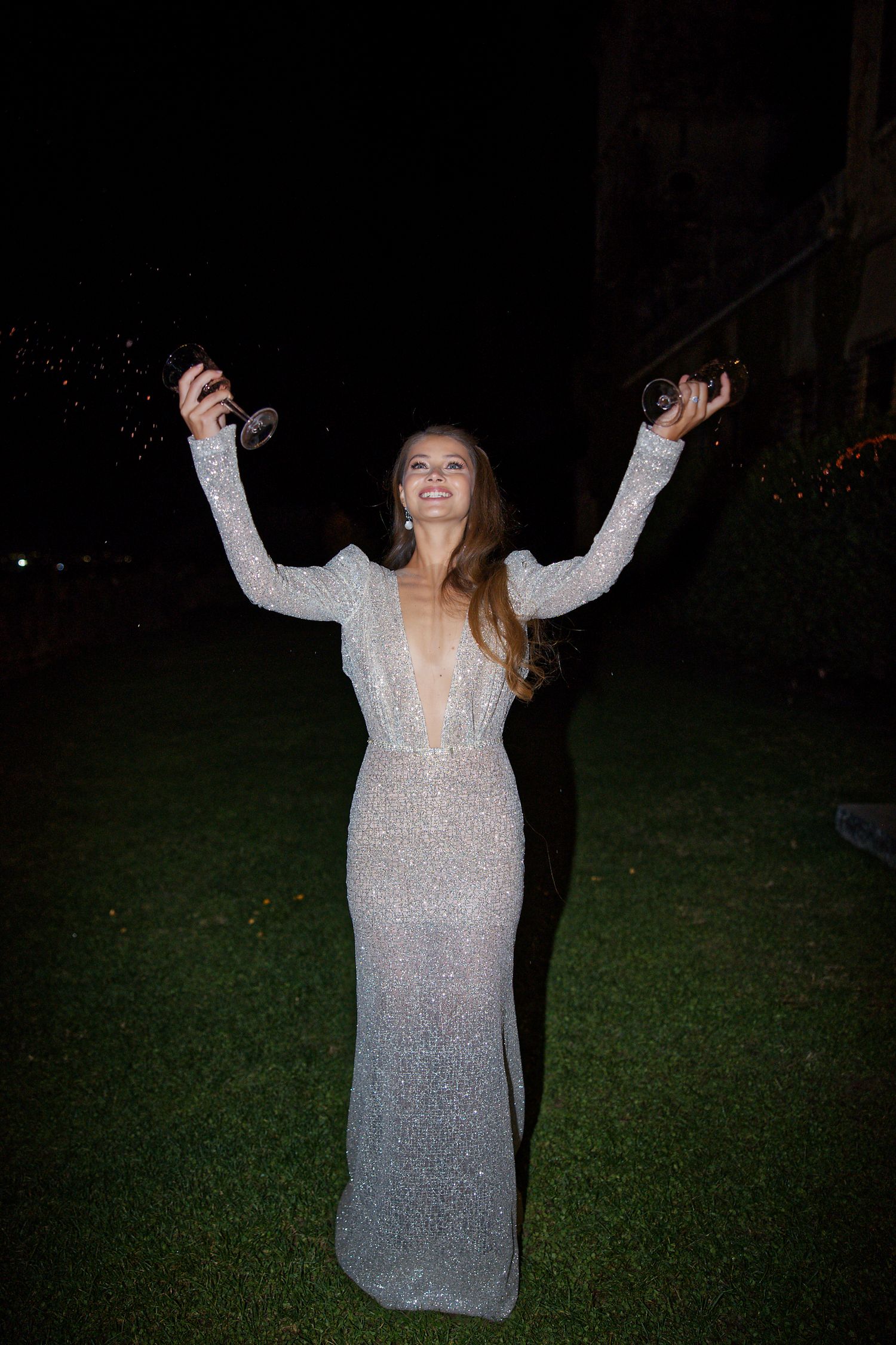 A person in a sparkling white evening gown poses with raised arms on a dark lawn at night.