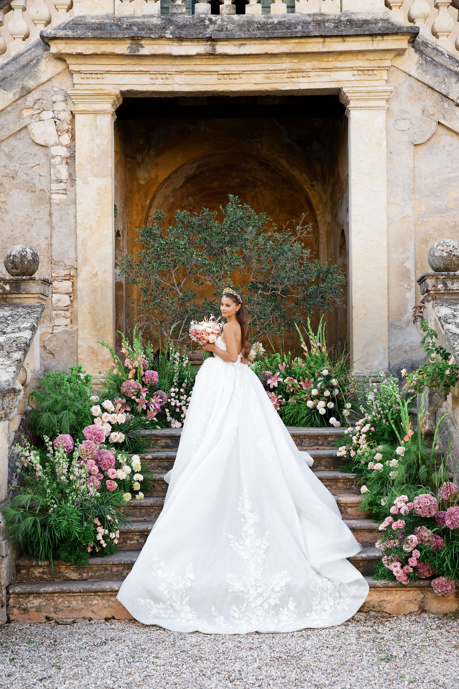 A bride in a flowing white gown with long train poses on stone steps surrounded by pink flowers and greenery.