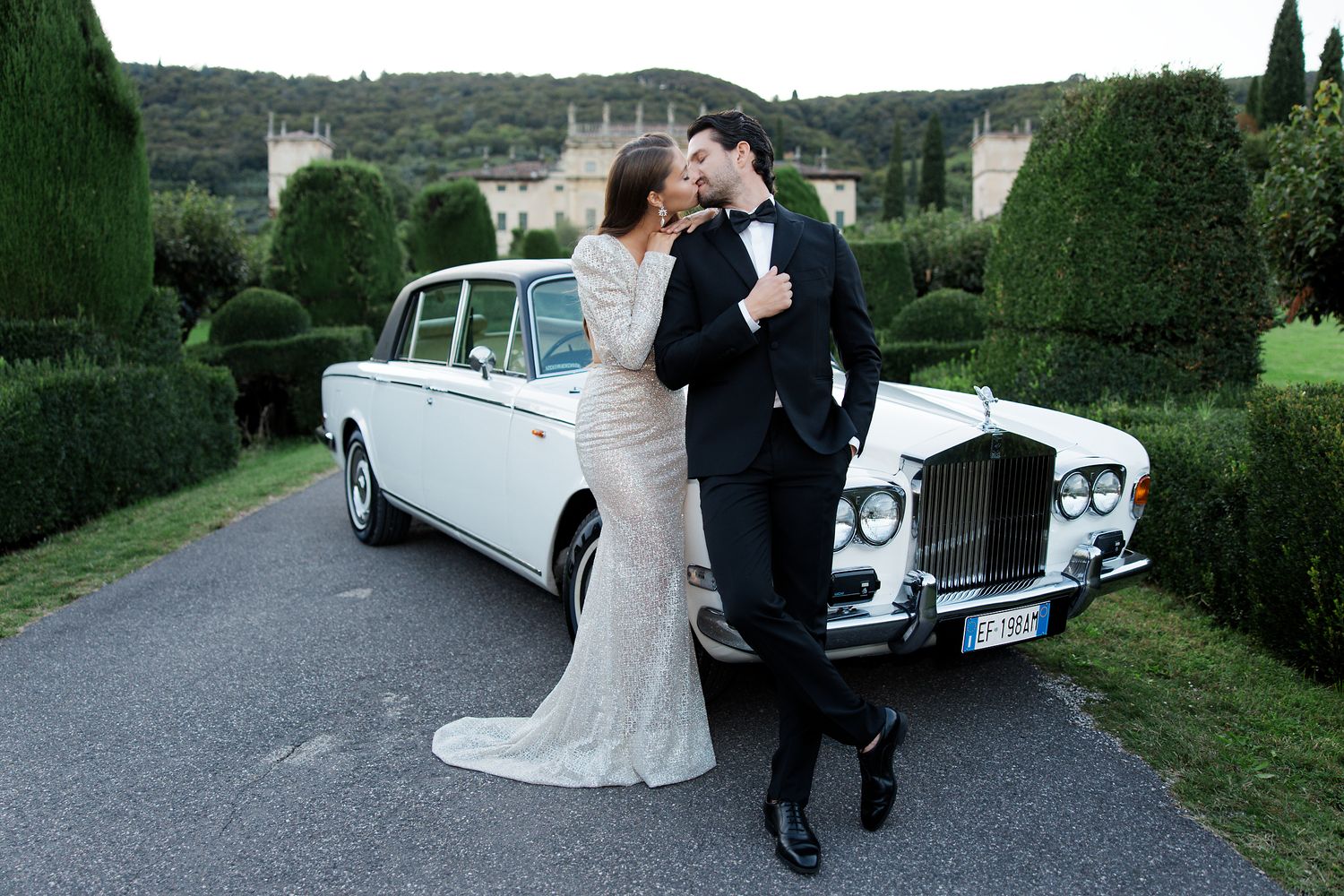 A couple celebrates by a classic white Rolls Royce on a manicured garden driveway.