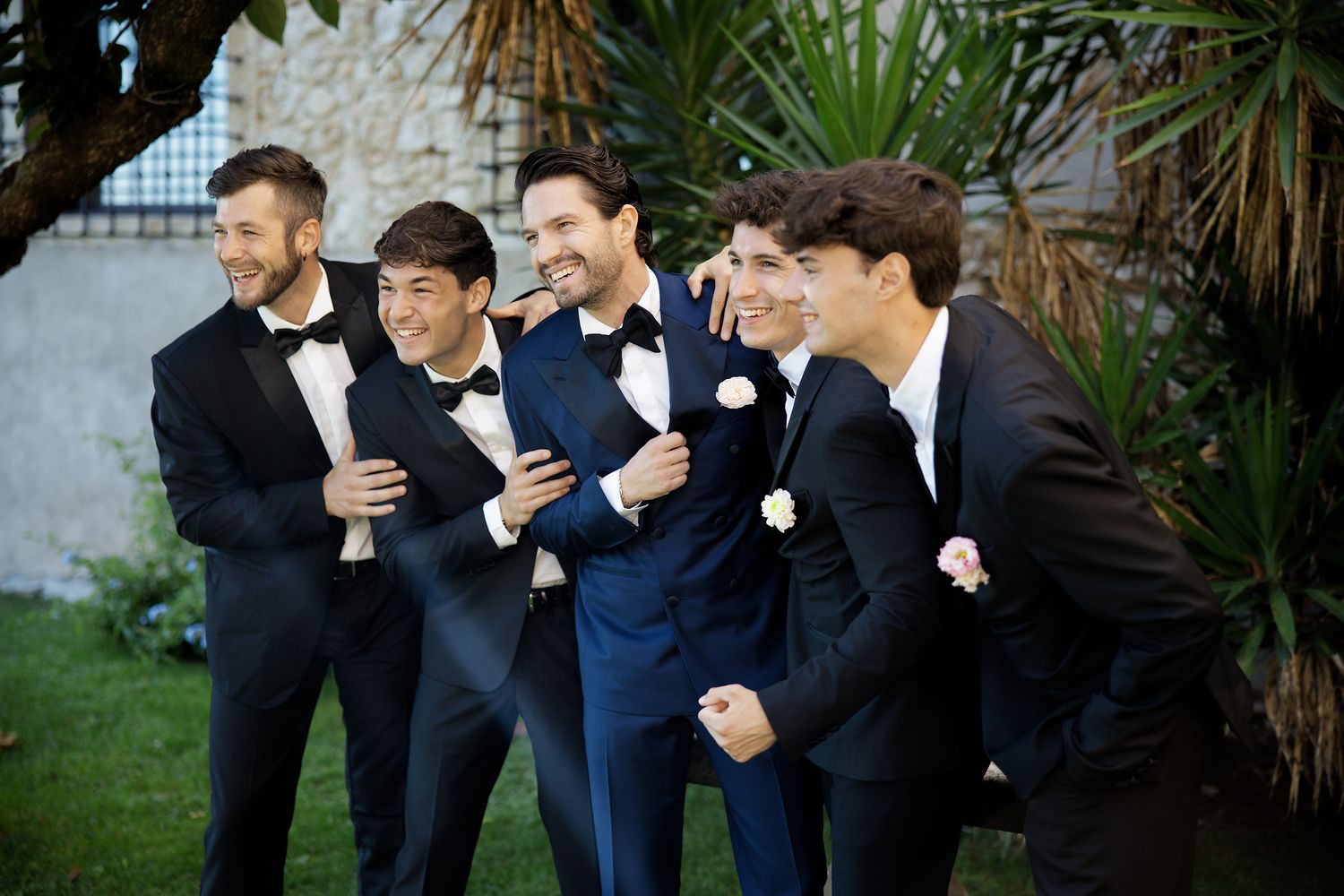 Groomsmen in black tuxedos share a candid moment of laughter outdoors.