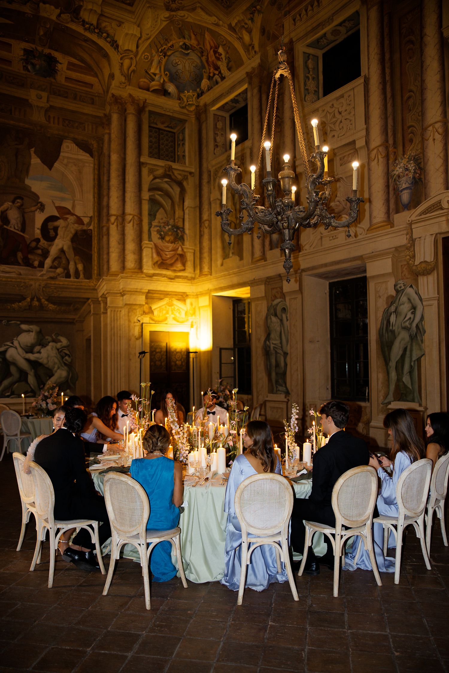 Elegant wedding reception in an ornate baroque hall with chandelier lighting and white chairs around dining tables.