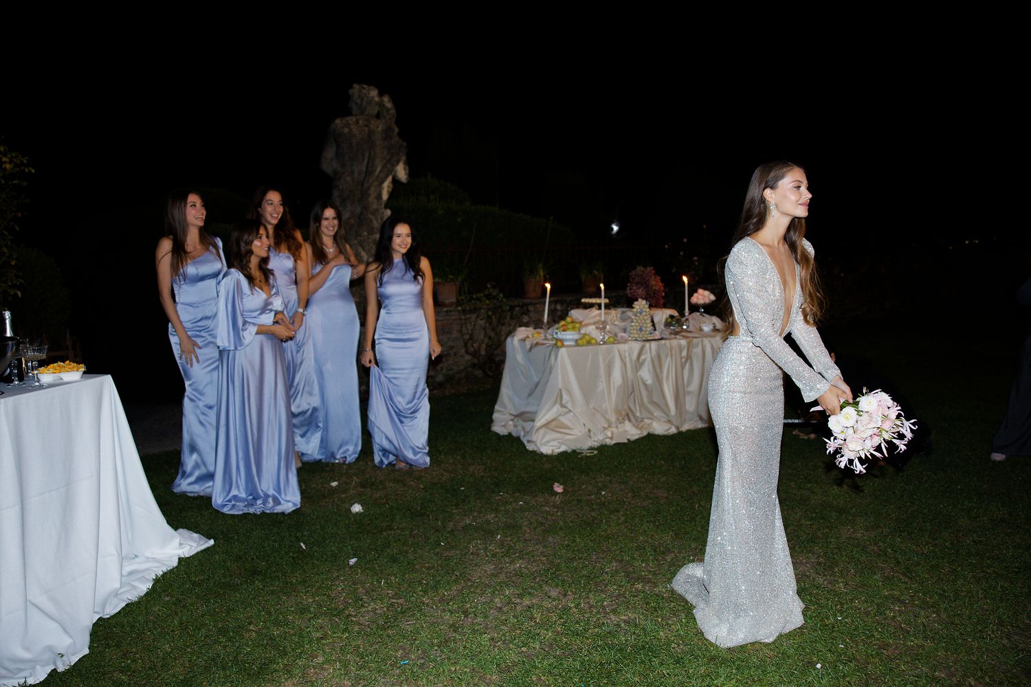 Wedding party scene with bridesmaids in blue dresses gathered around decorated reception tables at dusk.