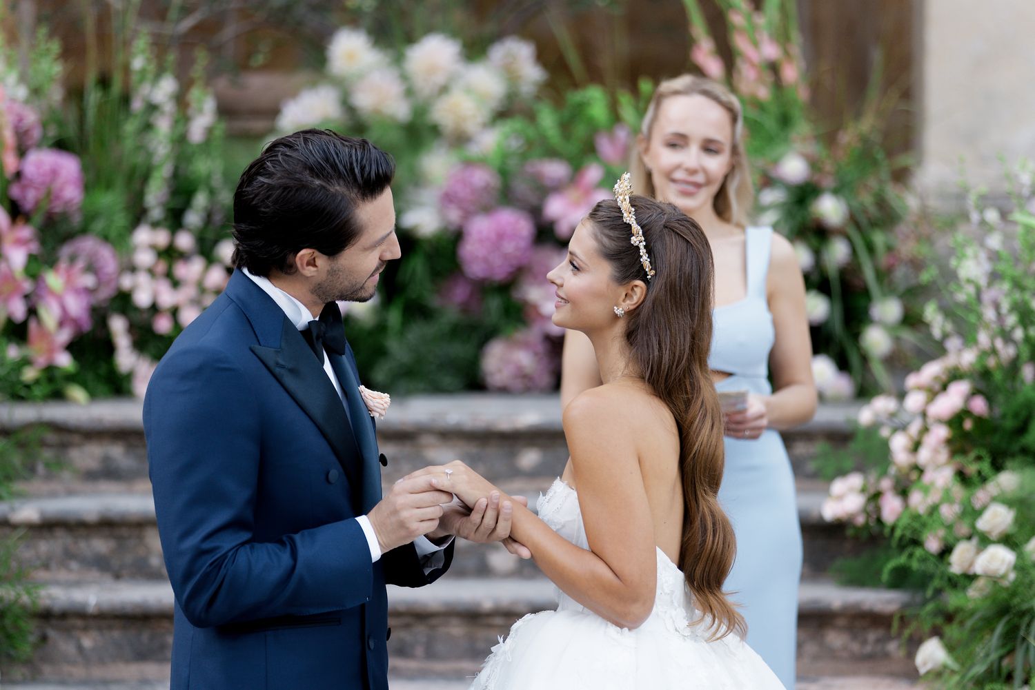 Romantic wedding scene in a garden with purple flowers and stone steps as backdrop.