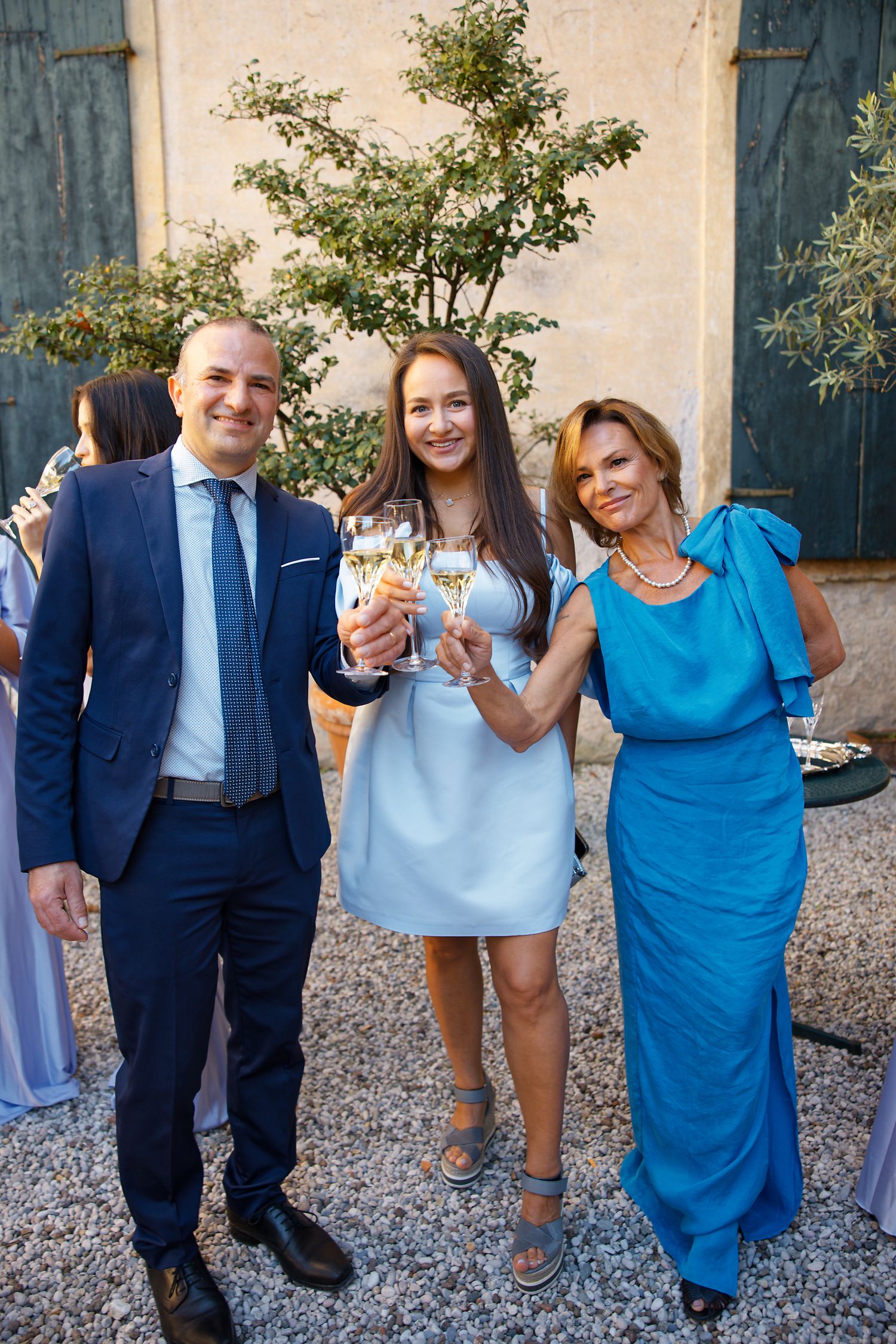 Three people dressed in formal attire pose with wine glasses at an elegant outdoor gathering.
