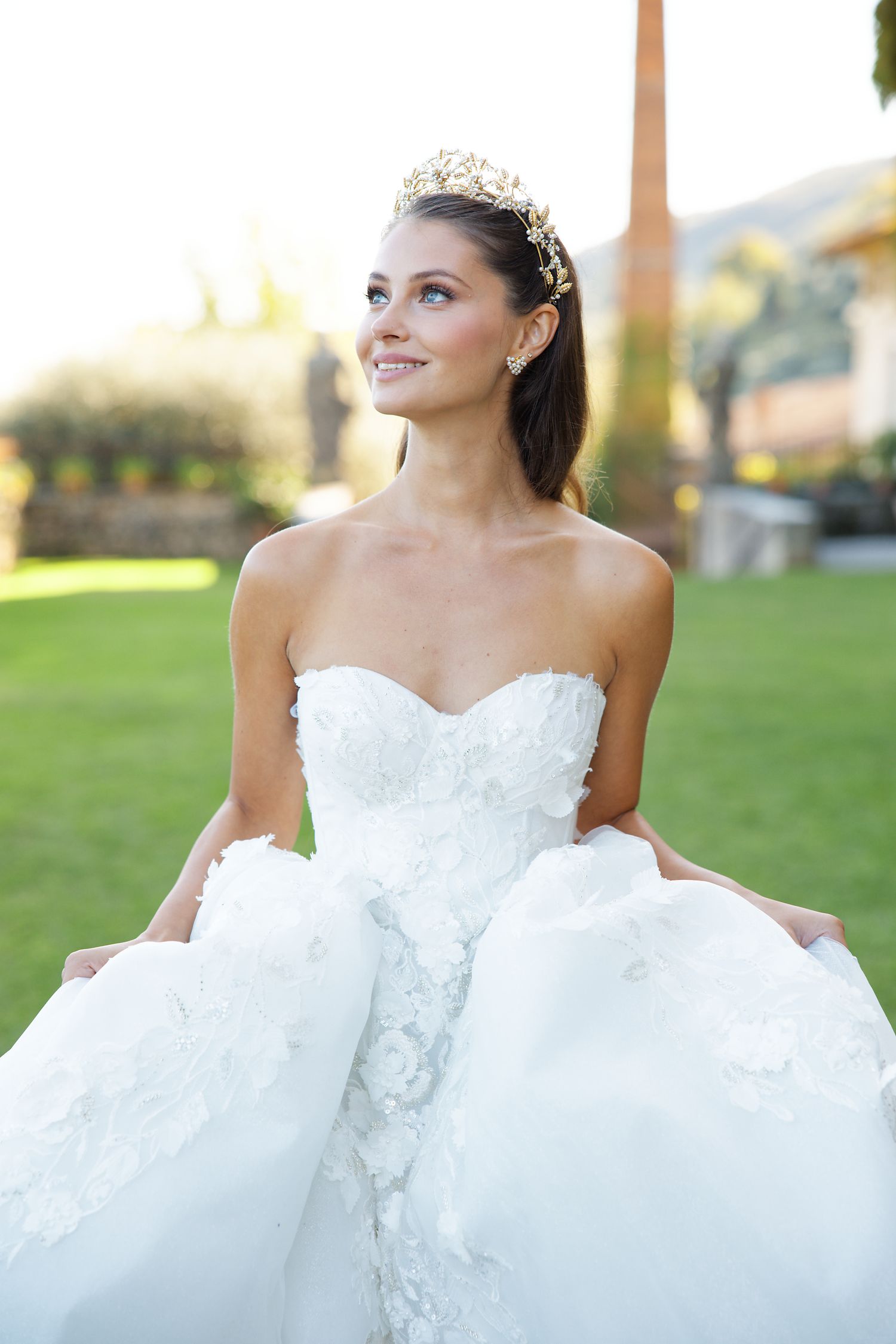 A bride in a white strapless ballgown and tiara poses outdoors in soft sunlight.