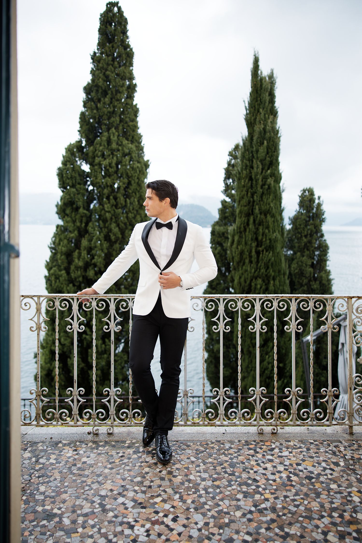 Stylish groom in white dinner jacket stands on Mediterranean terrace with lake view.