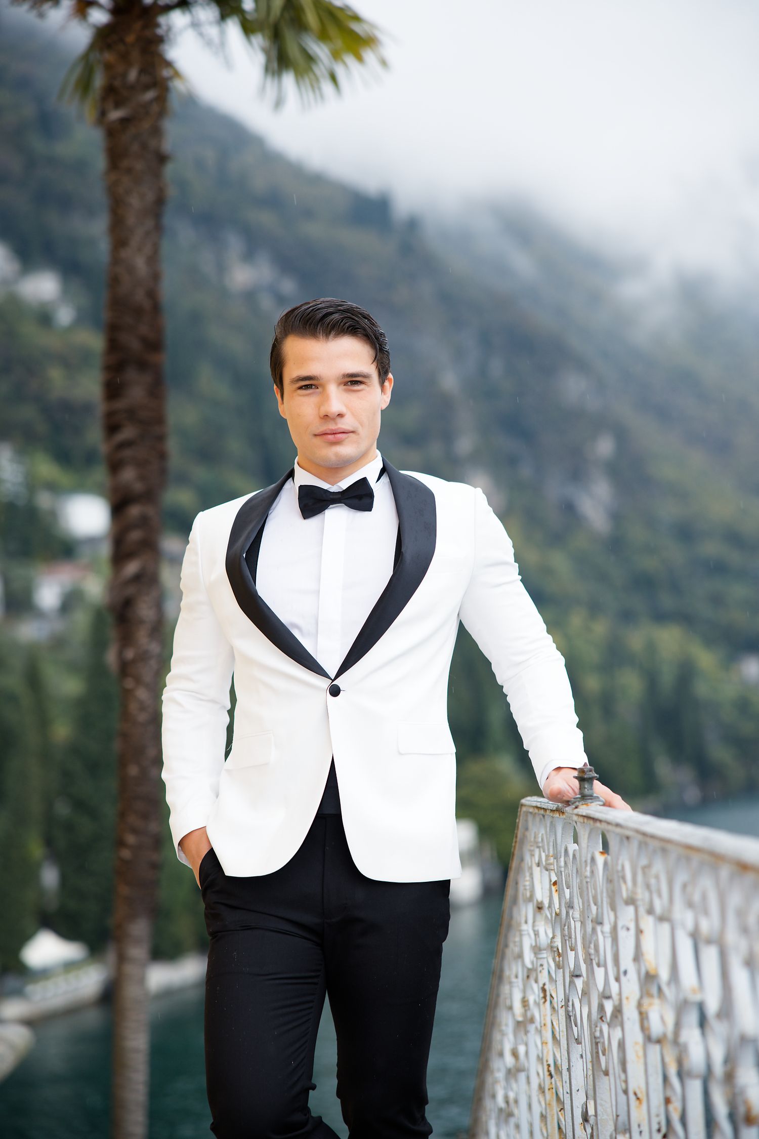 Groom in classic white dinner jacket stands on balcony with palm trees and mountain vista.