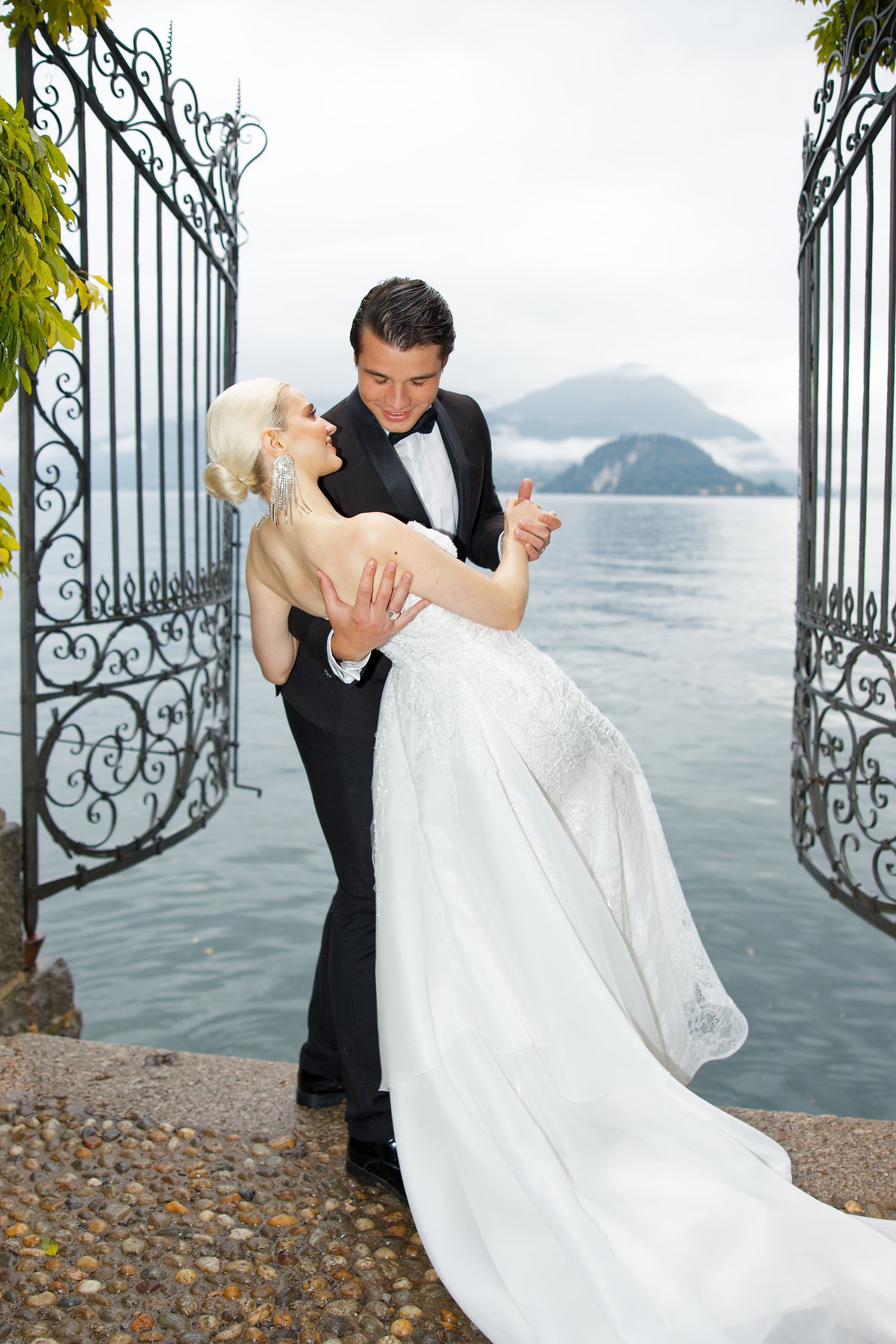 Romantic wedding photo with couple in formal attire embracing by ornate gates overlooking a misty lake and mountains.