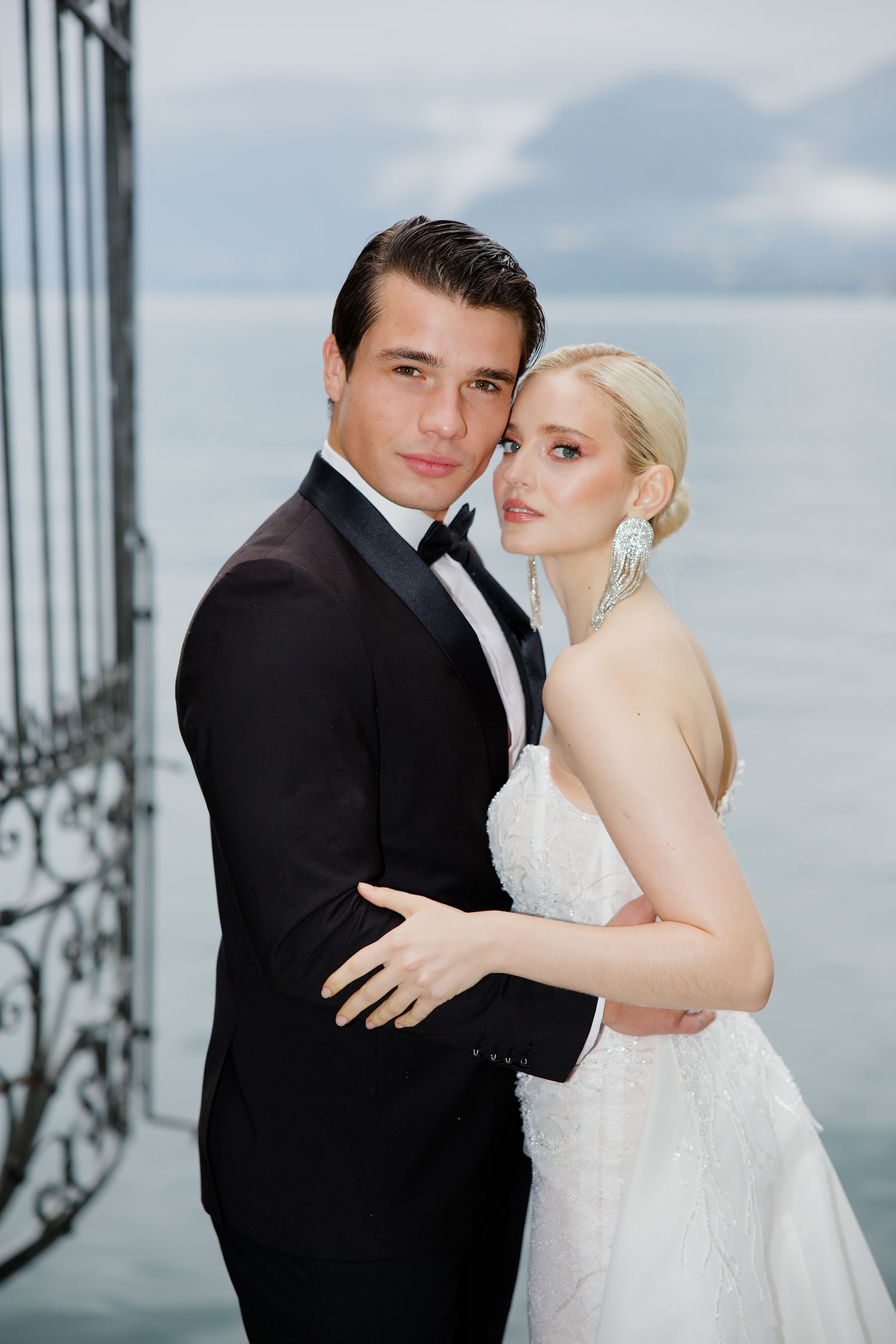 A formal couple in wedding attire pose against a lakeside backdrop.