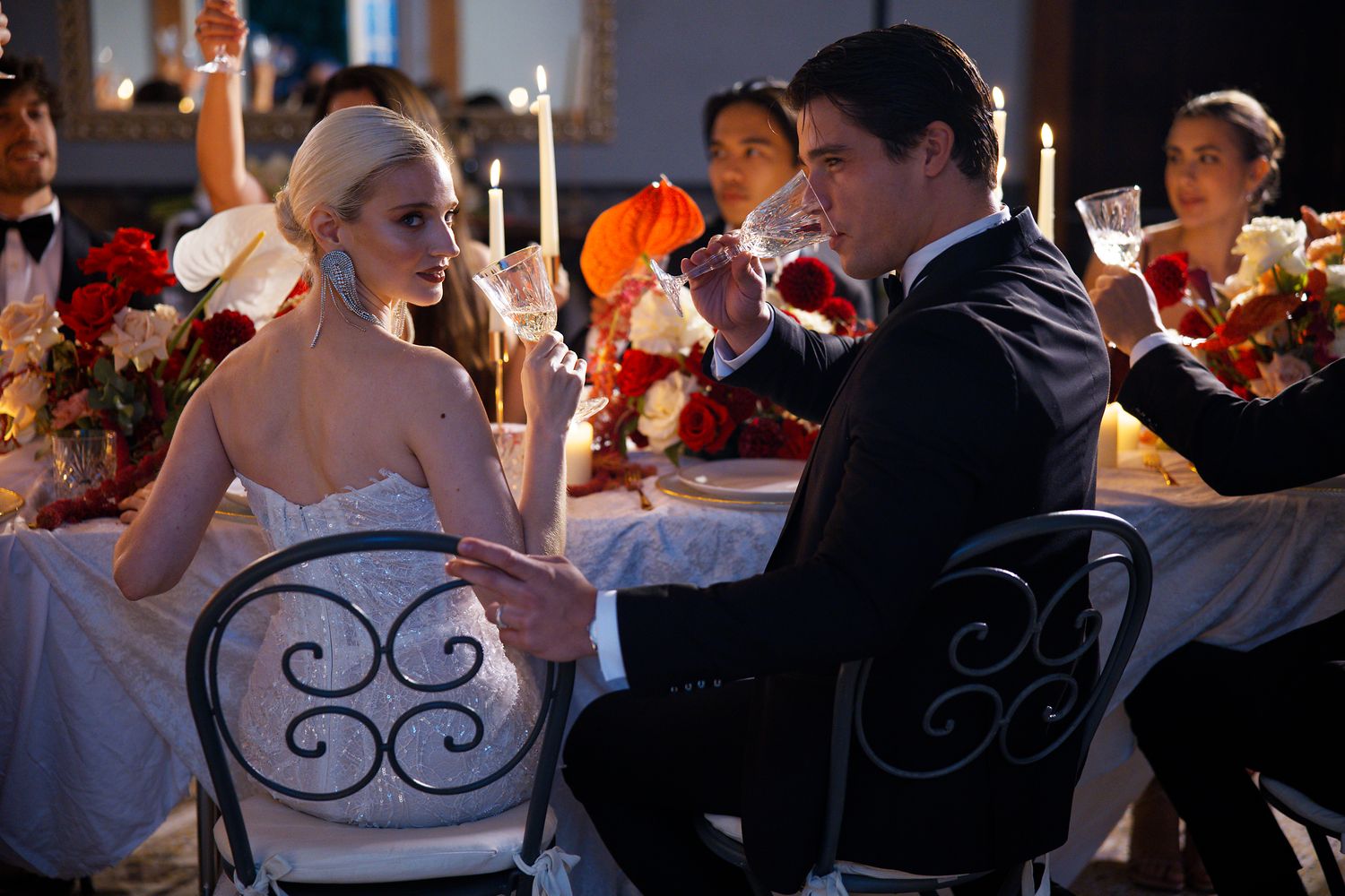 Wedding couple sits in ornate chairs at a candlelit reception table during their celebration.