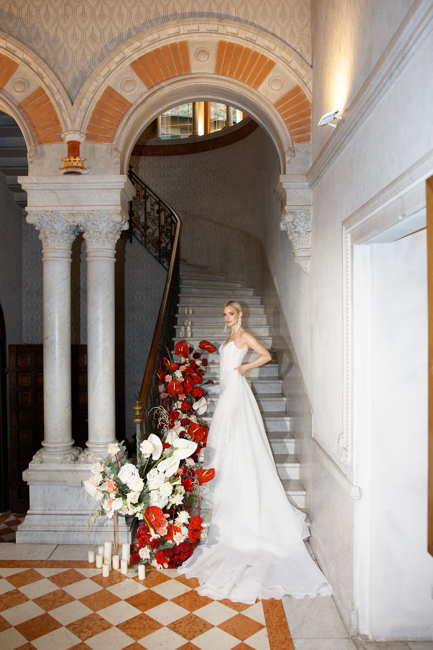 Bridal portrait in classic venue featuring arched columns and dramatic red and white floral staircase decor.