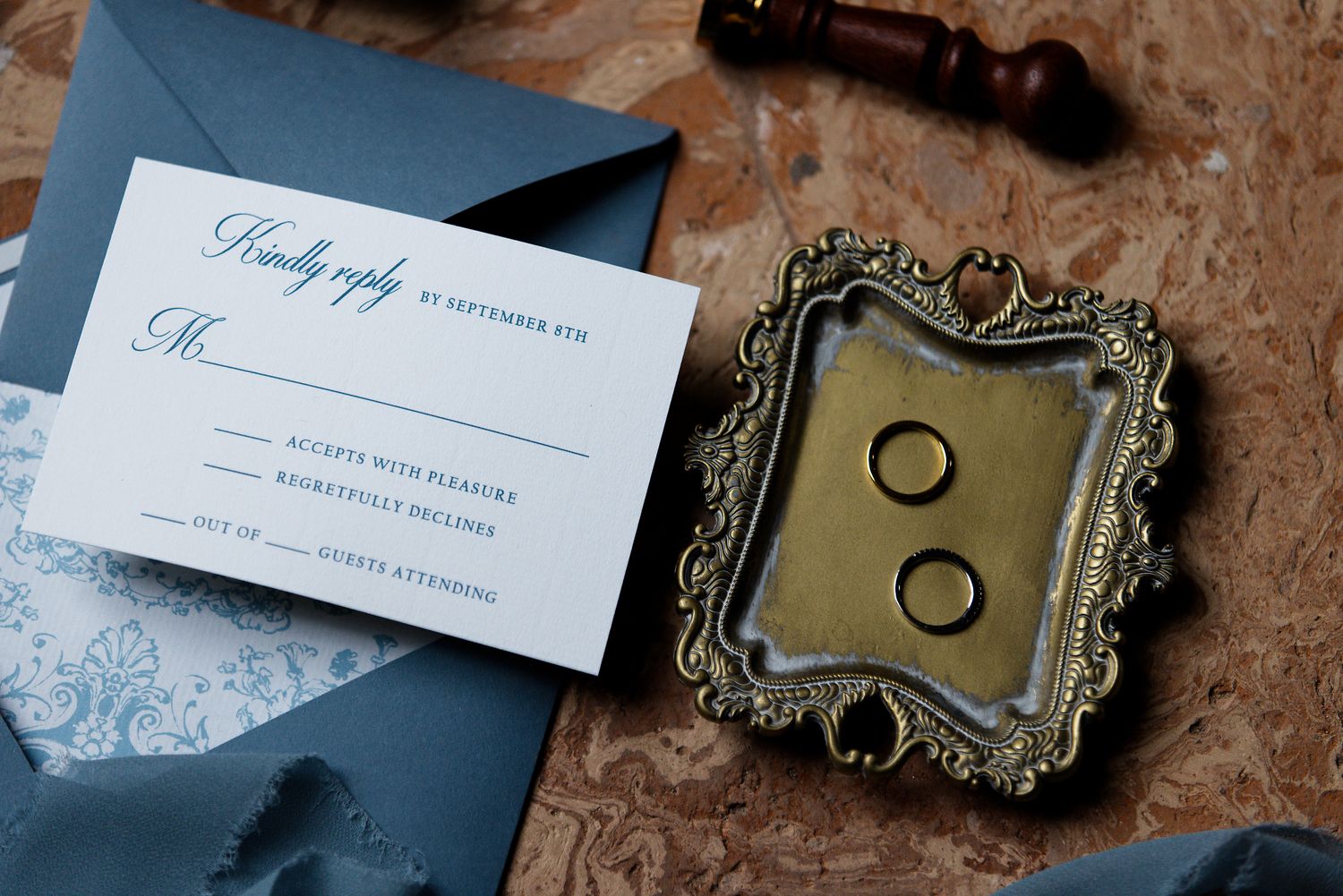 Wedding invitation and vintage ornate frame with rings displayed on wooden surface.