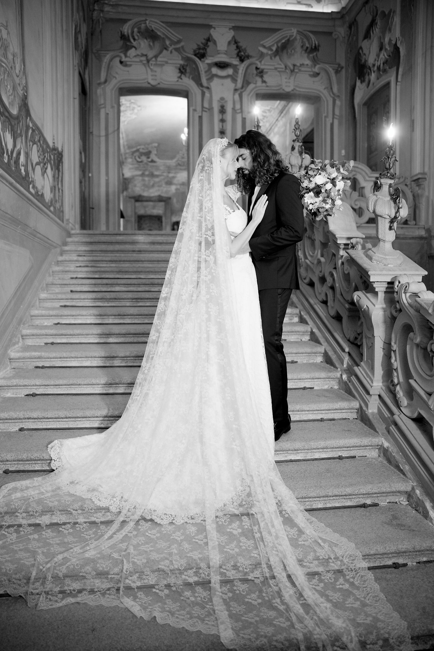A romantic black and white wedding photo showing a couple embracing on an ornate marble staircase with a long lace veil.