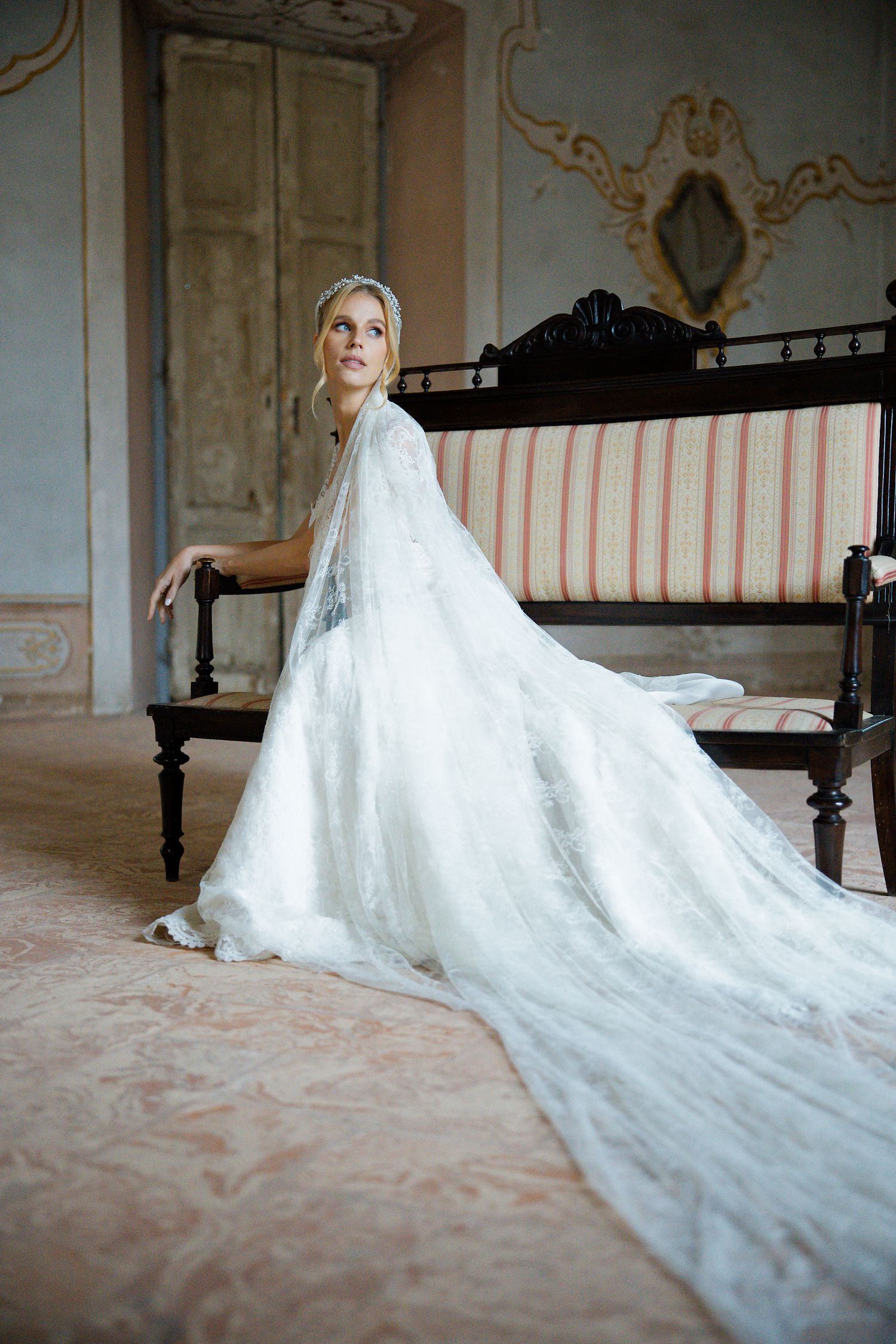 A flowing white wedding gown with a long train drapes elegantly across an antique settee in an ornate room.