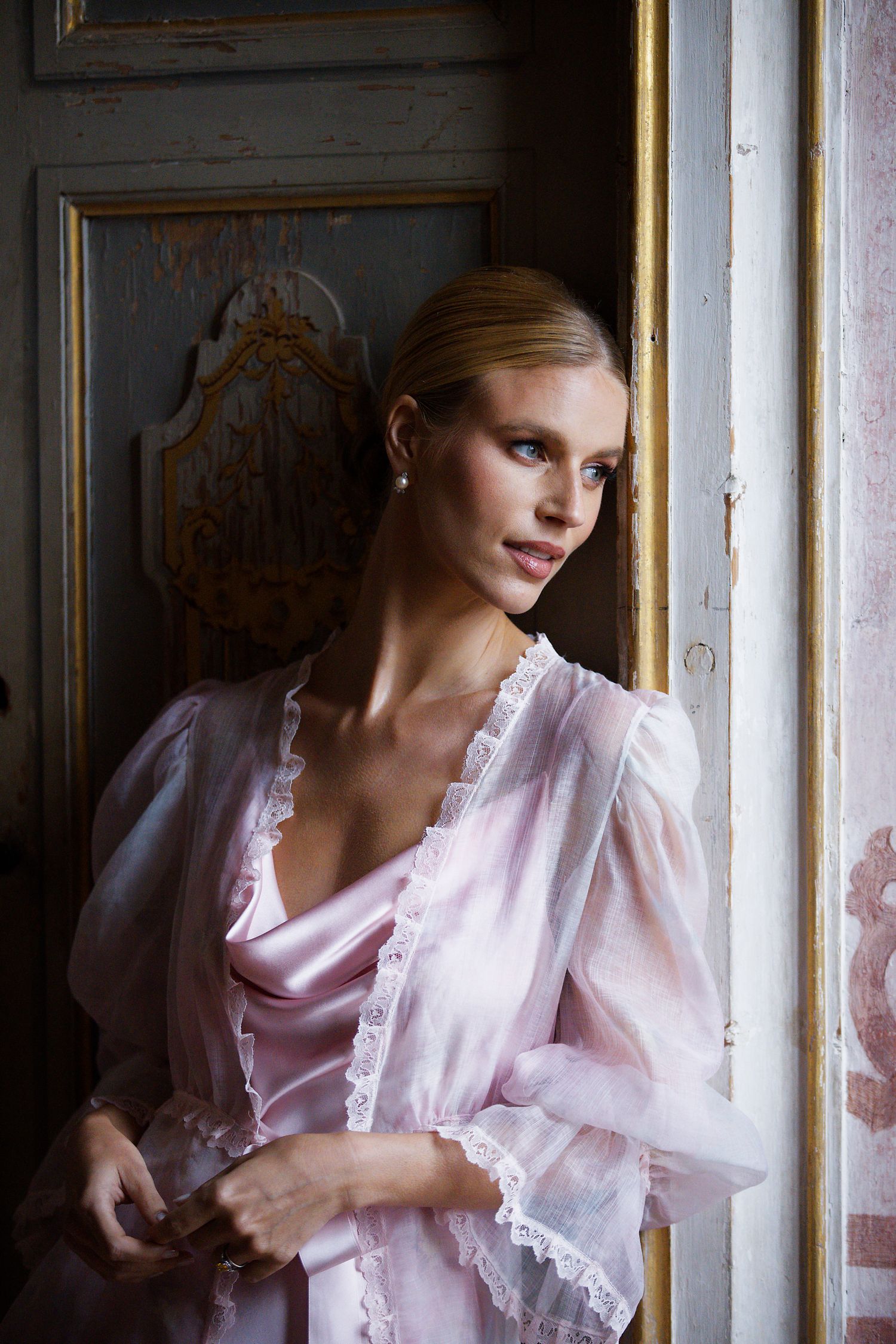 A figure in a delicate pink vintage-style dress with lace trim stands near a window in moody lighting.
