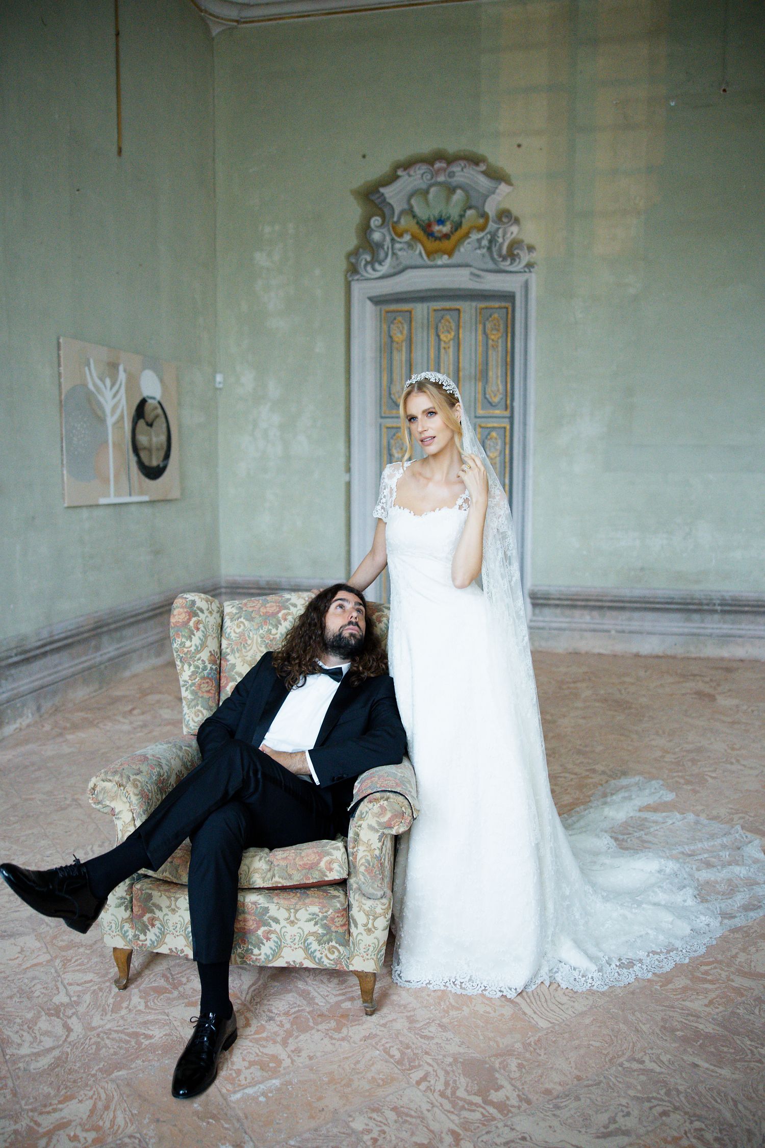 Couple poses in vintage room with faded green walls, person in dark suit lounging on floral armchair next to figure in white gown.