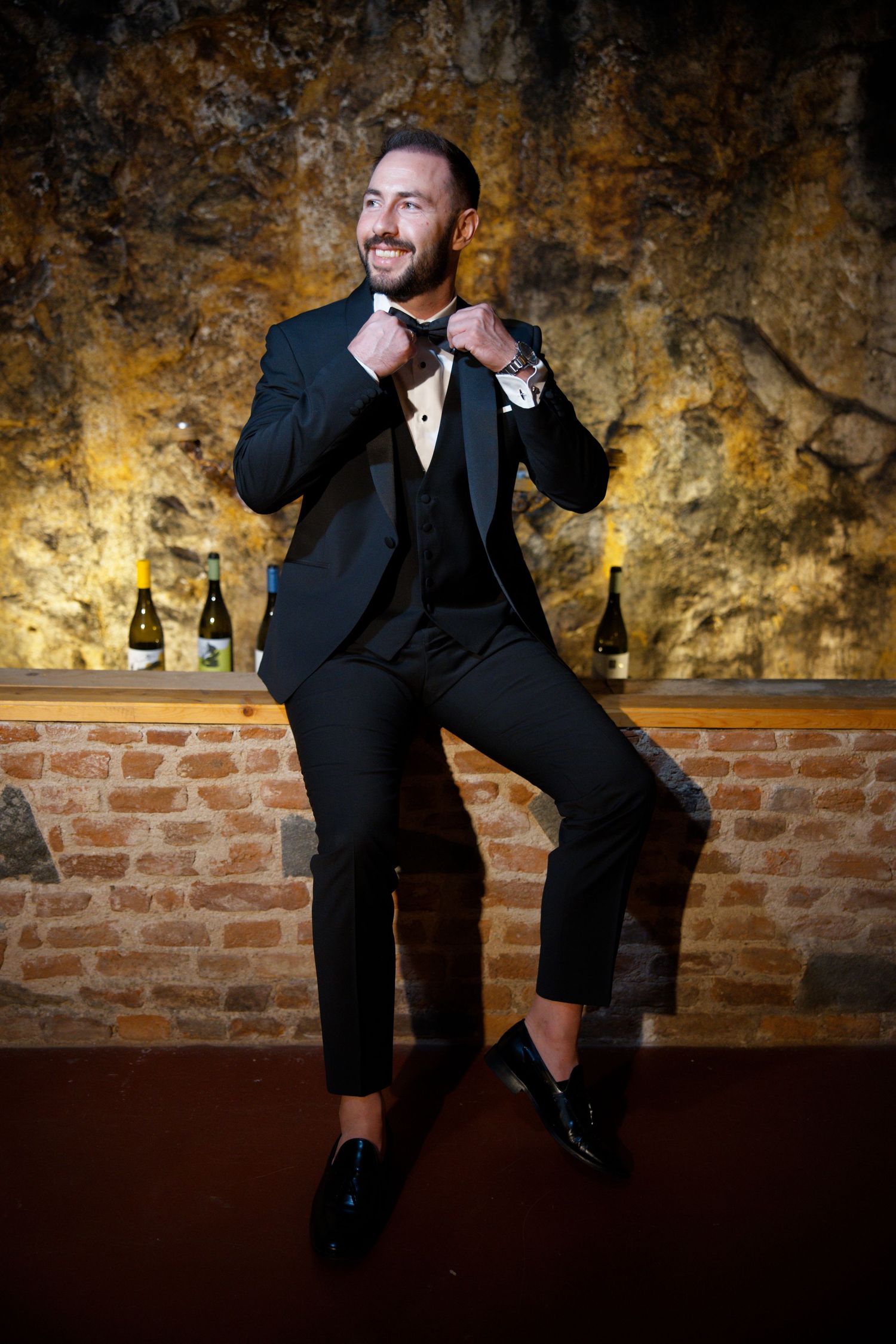 A well-dressed figure in a black tuxedo adjusts their bowtie against a textured golden wall backdrop.