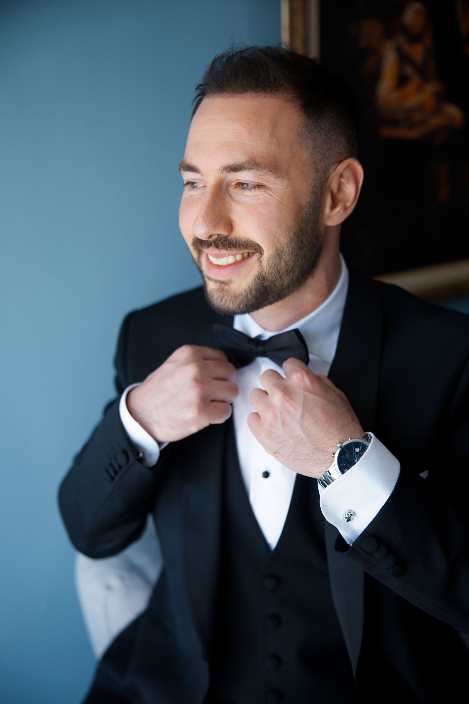 Stylish groom in black tuxedo adjusts his bowtie while getting ready for the wedding.
