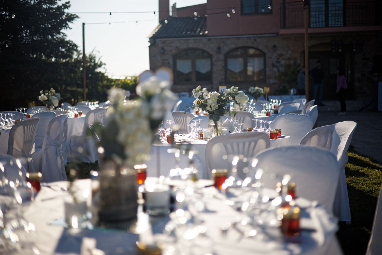 Outdoor wedding reception setup with white chairs and floral centerpieces at sunset.