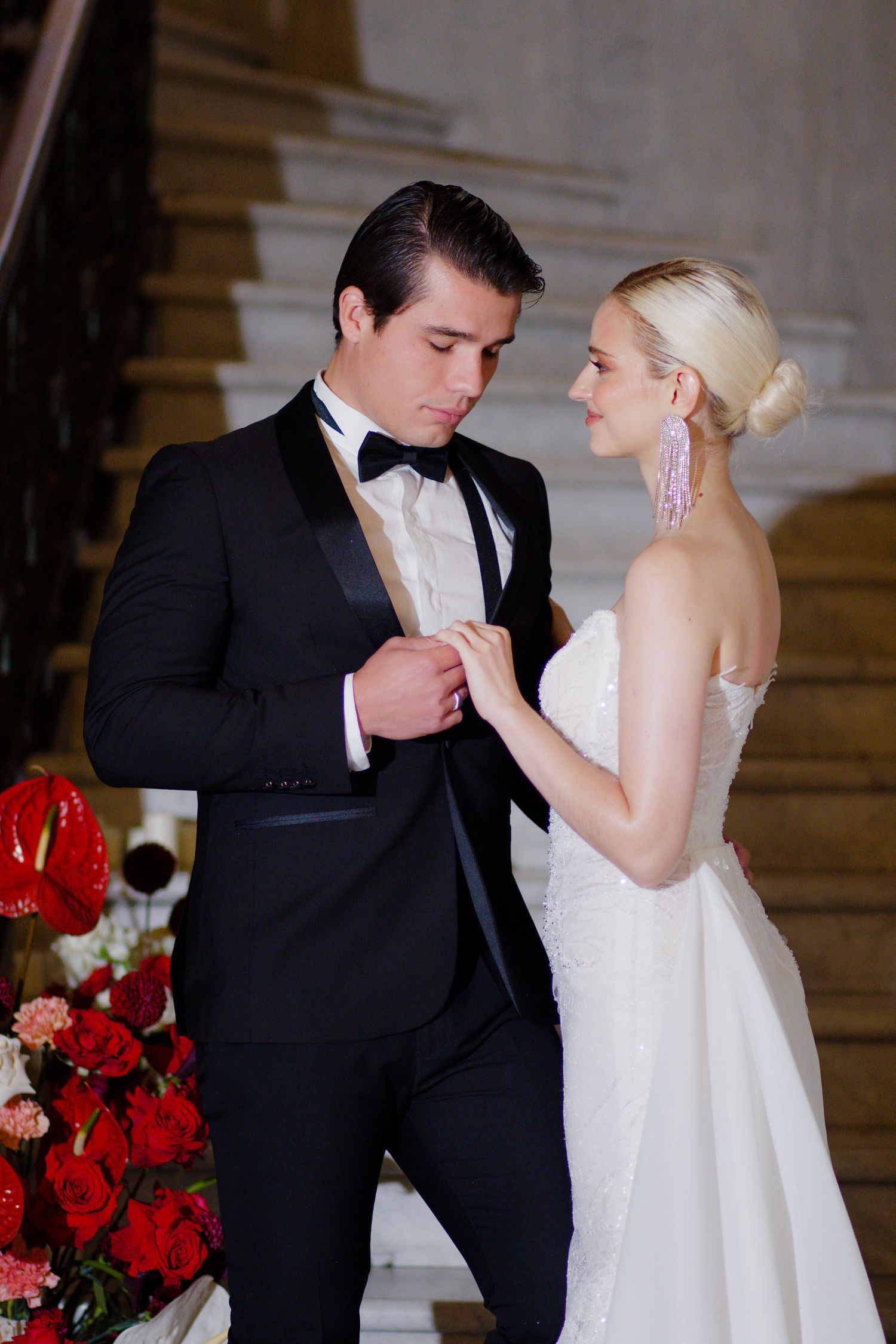 A couple shares an intimate moment on a grand staircase with red floral decorations.