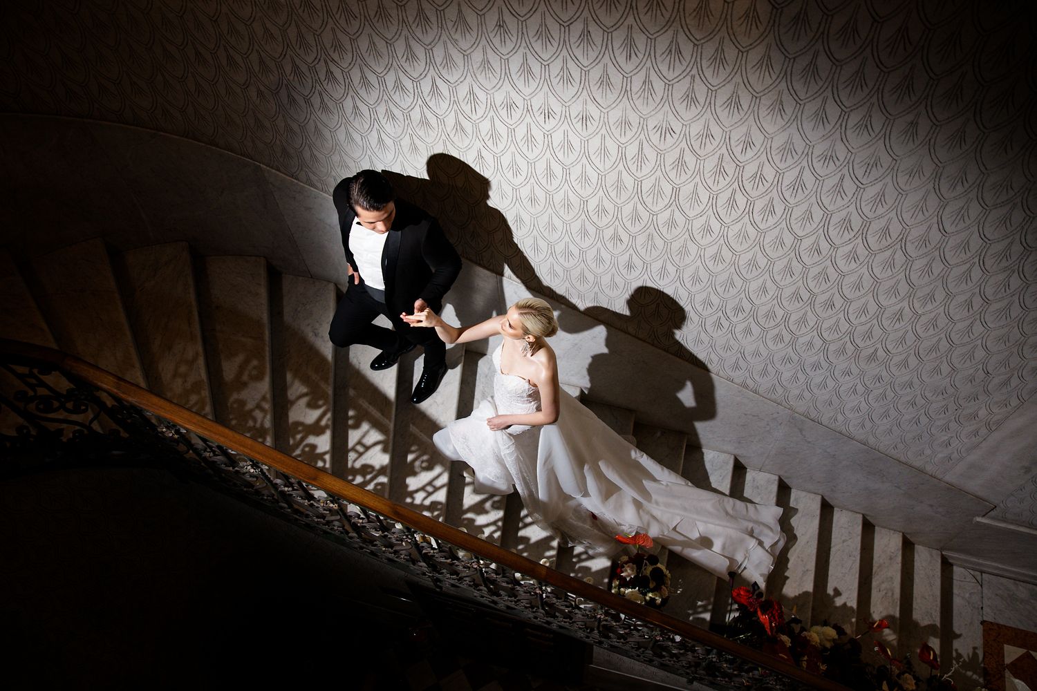 Dramatic wedding photo on staircase with artistic shadows cast on patterned wallpaper.