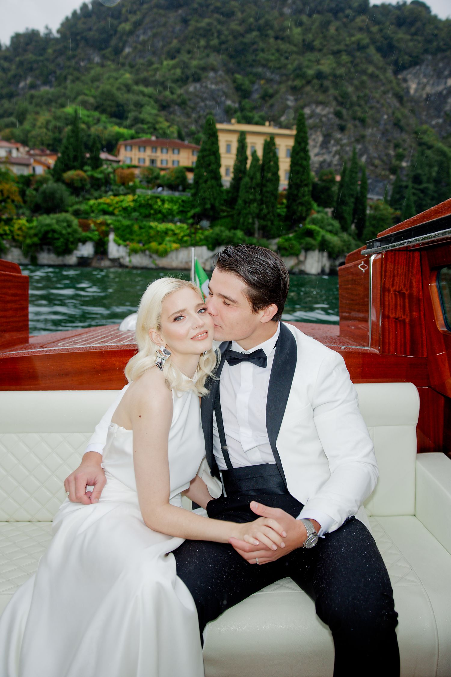 Couple shares tender moment on vintage wooden boat with Mediterranean villa scenery.