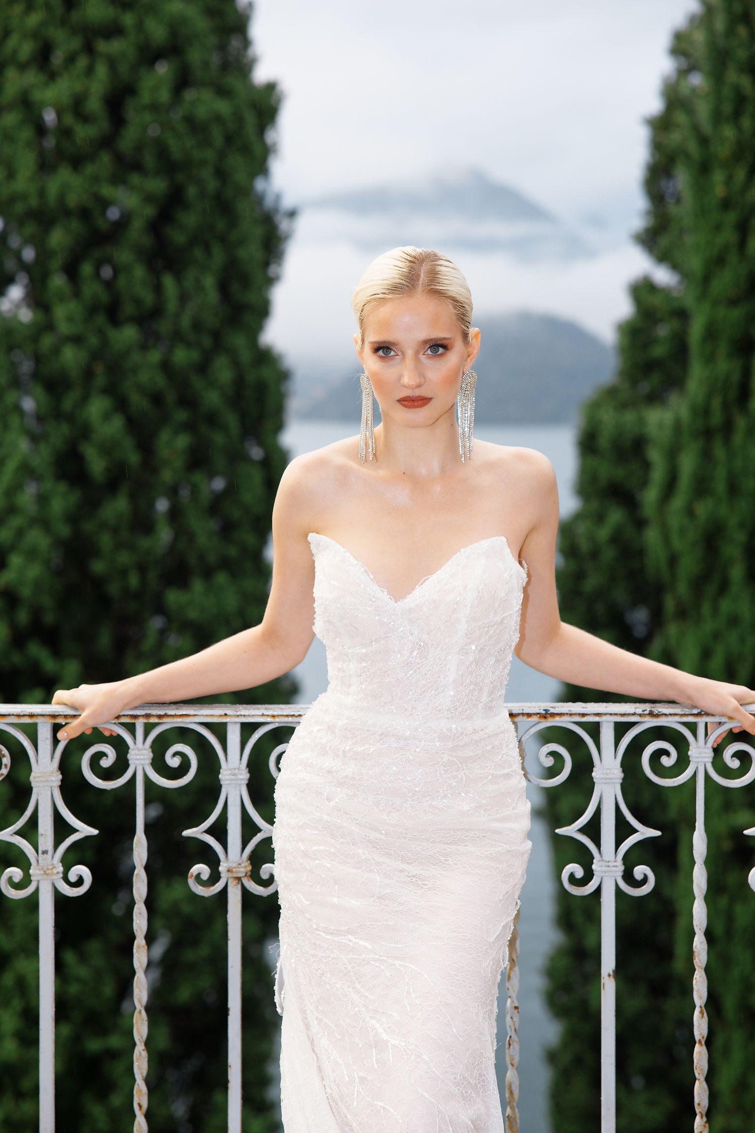 A bride in a strapless white gown poses on a balcony with cypress trees and mountains in the background.