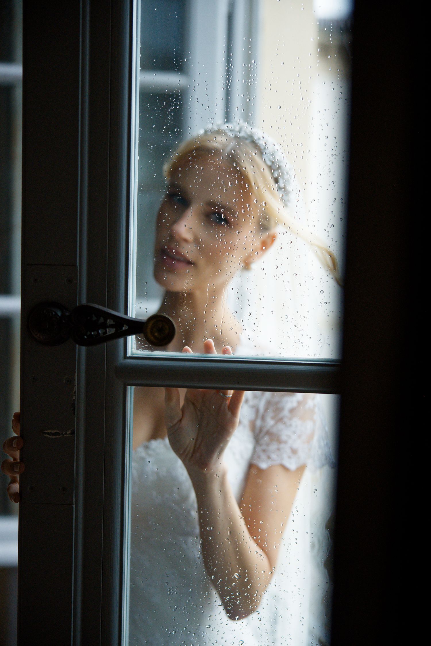 Artistic portrait through rain-spotted window glass in soft natural light.