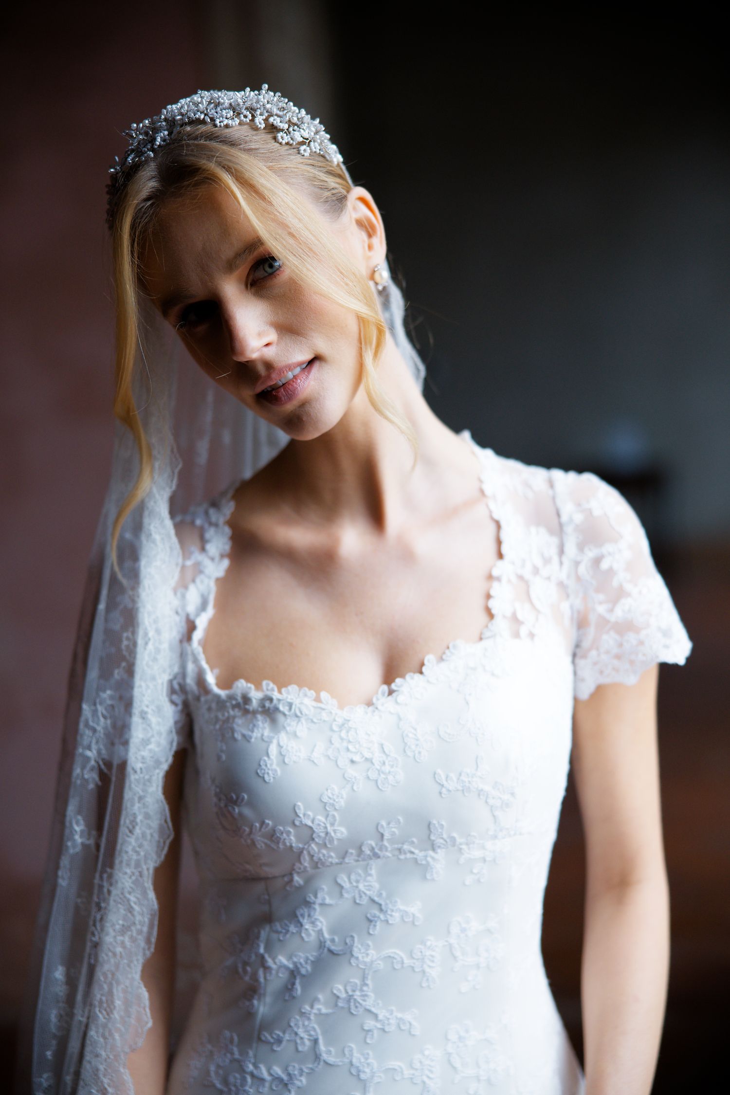 Bride wearing a delicate lace wedding dress with short sleeves and crystal tiara poses indoors.