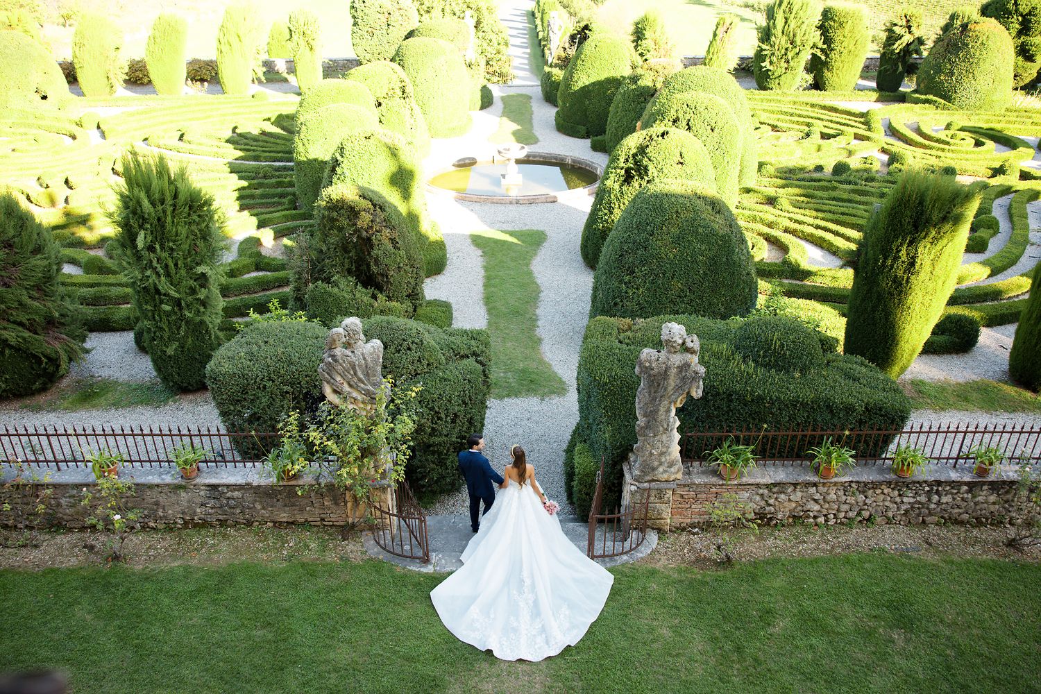 Aerial view of a wedding couple on manicured garden grounds with symmetrical hedges and stone statues.