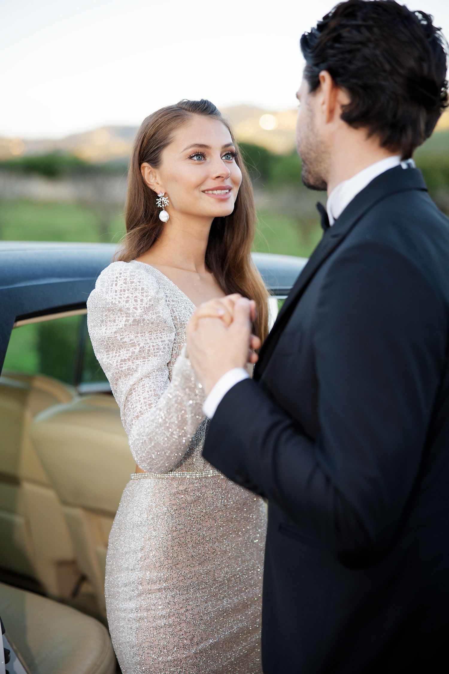 Couple in formal attire share an intimate moment beside a luxury car at sunset.
