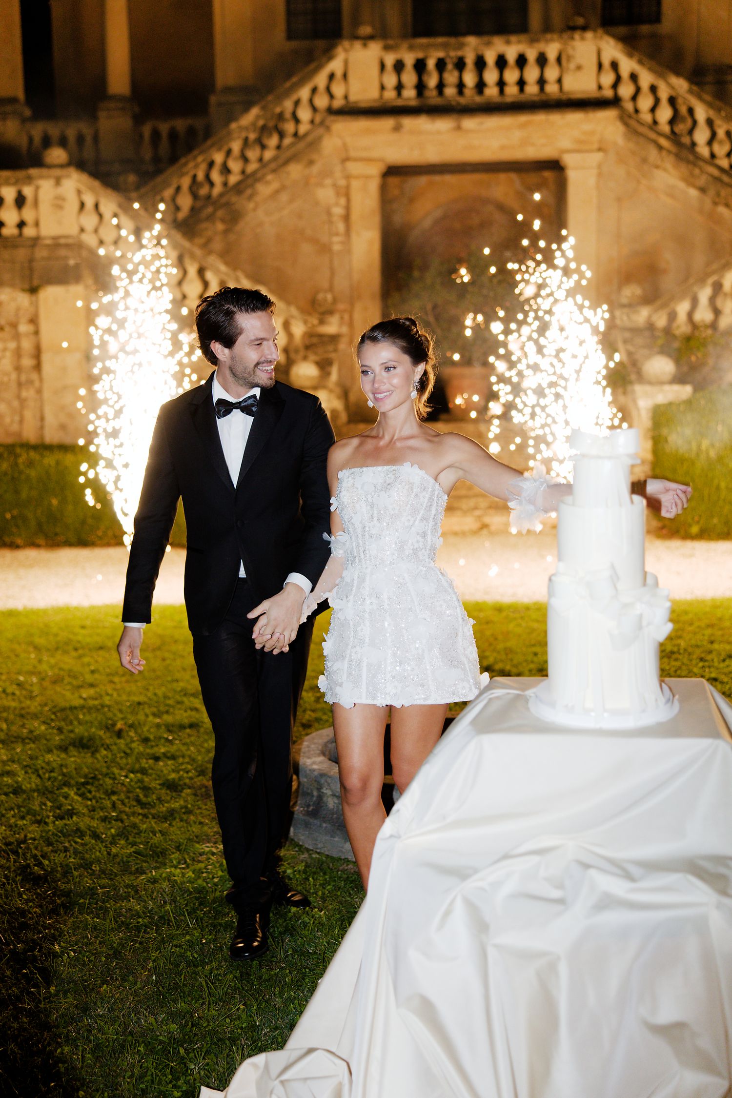 Couple walks past their wedding cake while sparklers illuminate the evening at a grand estate.