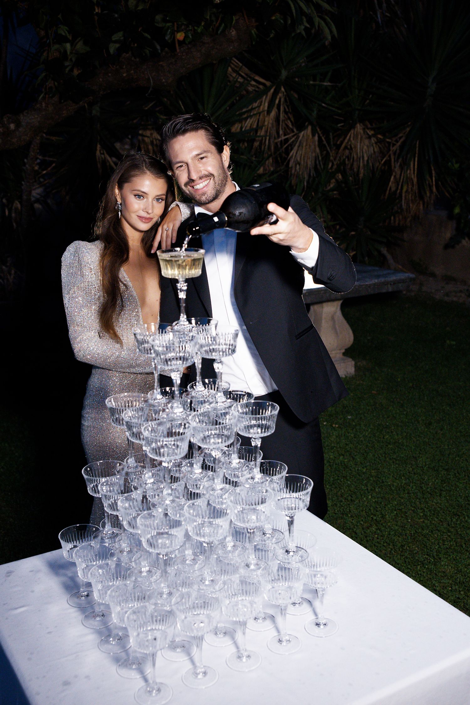 Elegant couple pouring champagne into tower of glasses at evening reception.