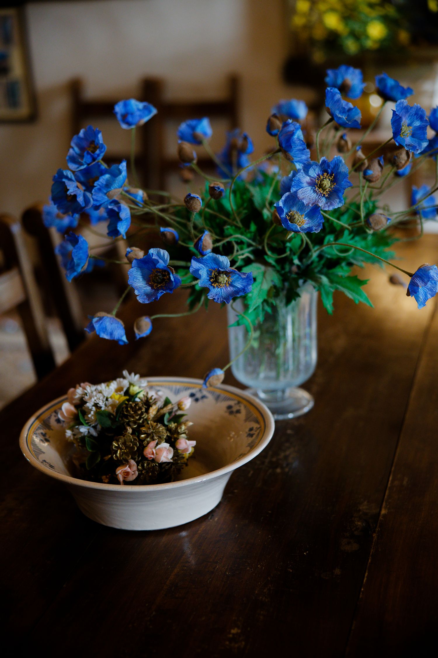 Vibrant blue flowers in a glass vase beside a ceramic bowl with small floral arrangement on dark wooden table.