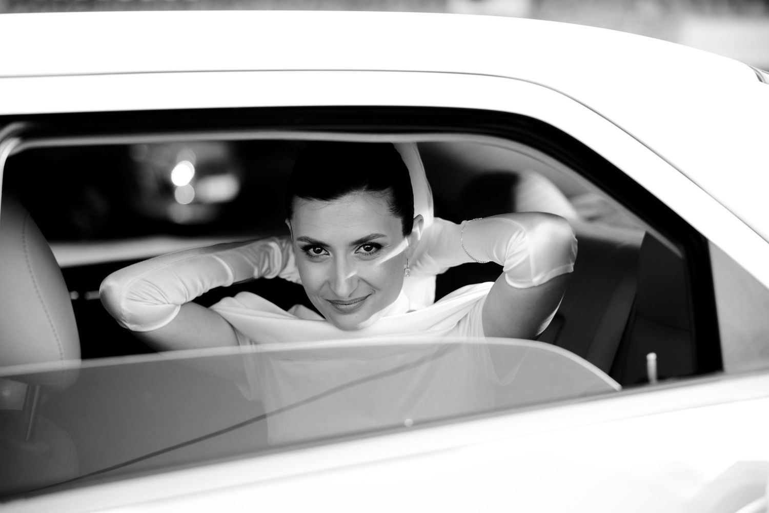 Black and white photo of someone looking playfully through a car window in an artistic portrait shot.