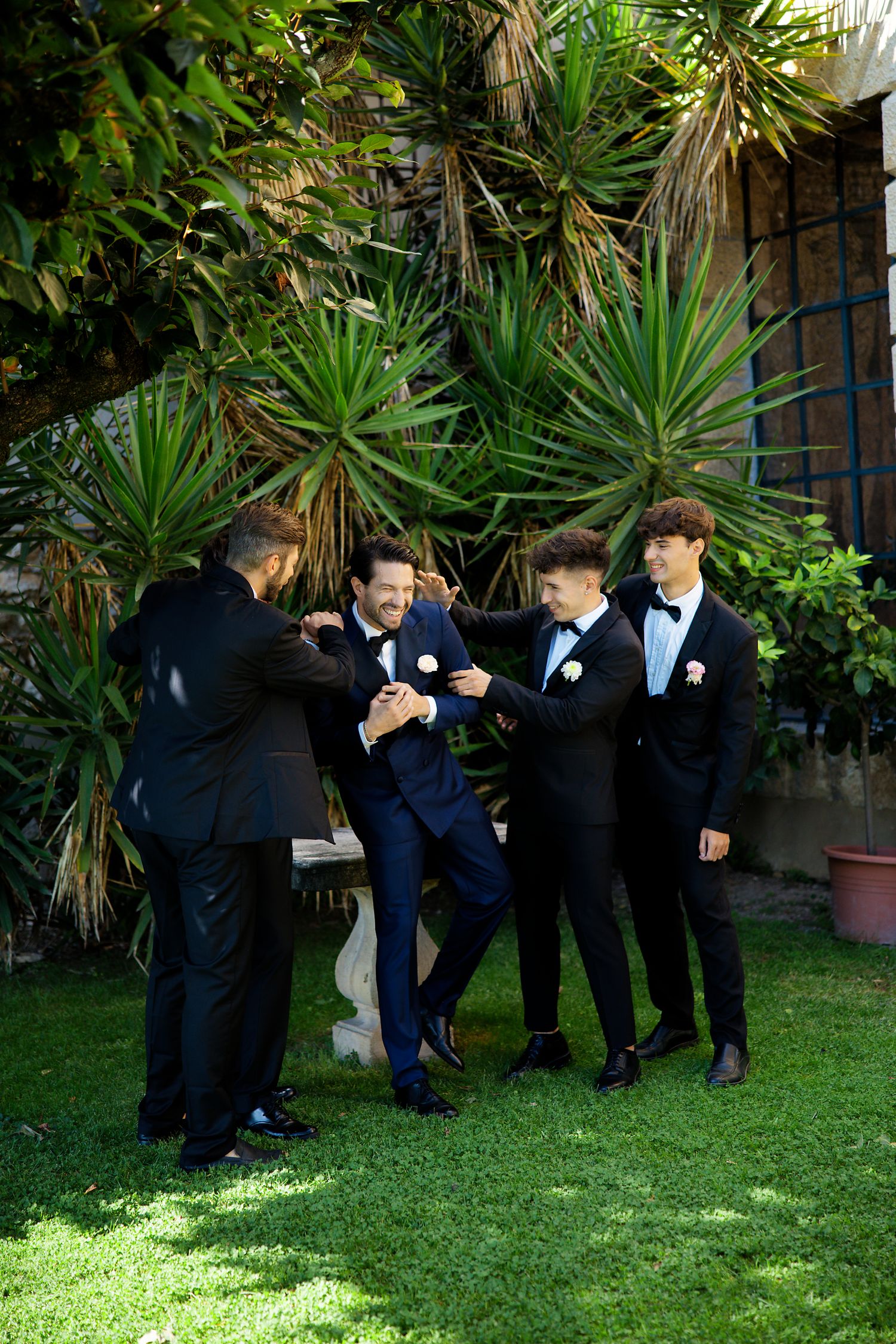 Group wearing formal black and navy suits gathered outdoors near tropical plants on a lawn.