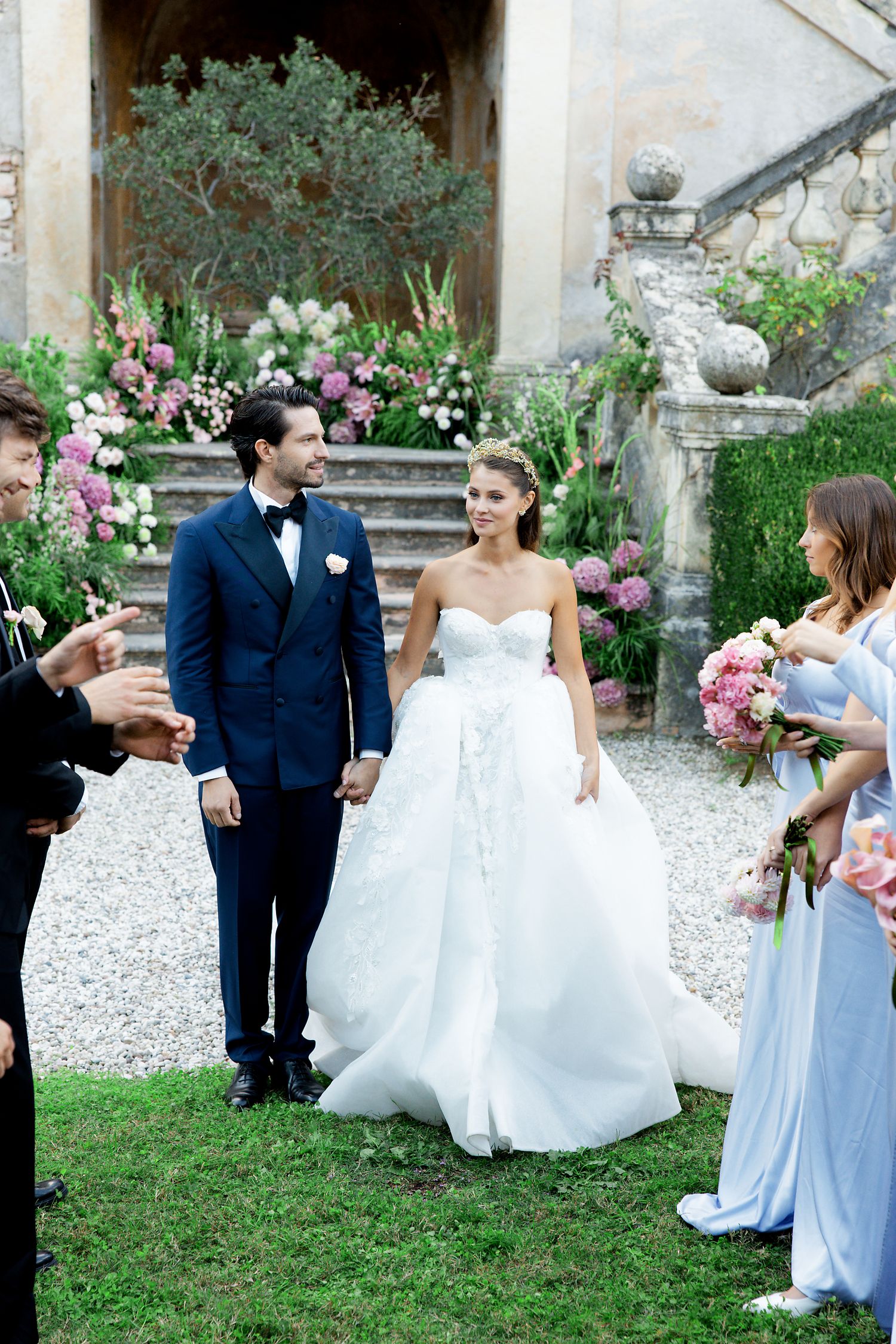 A wedding couple stands on stone steps surrounded by beautiful floral arrangements and guests at an elegant villa.
