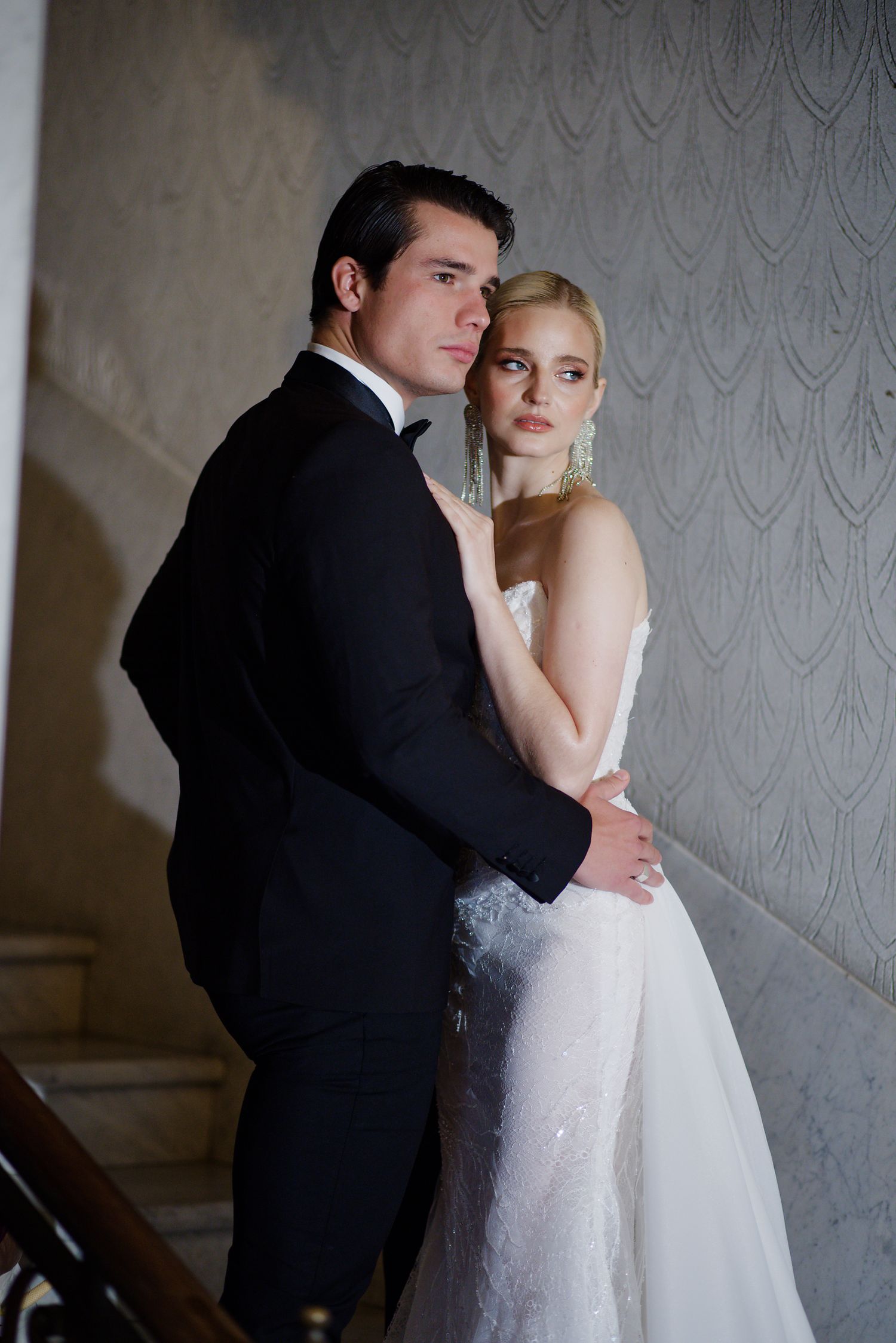 A romantic wedding portrait shows a couple in formal attire embracing against an elegant gray backdrop.