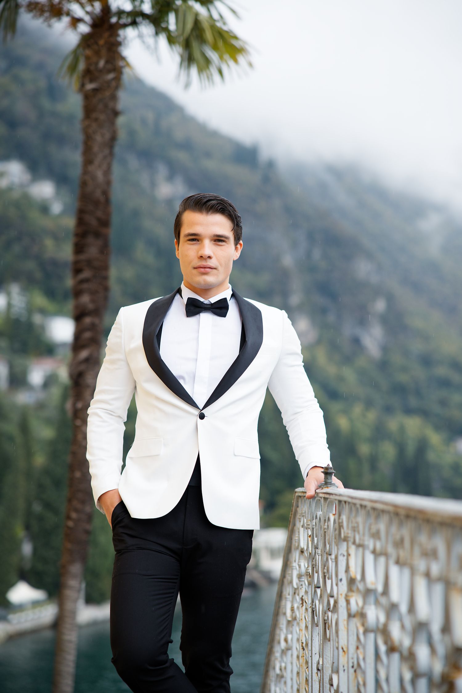 A person in a white dinner jacket and bow tie poses on a balcony with misty mountains in the background.