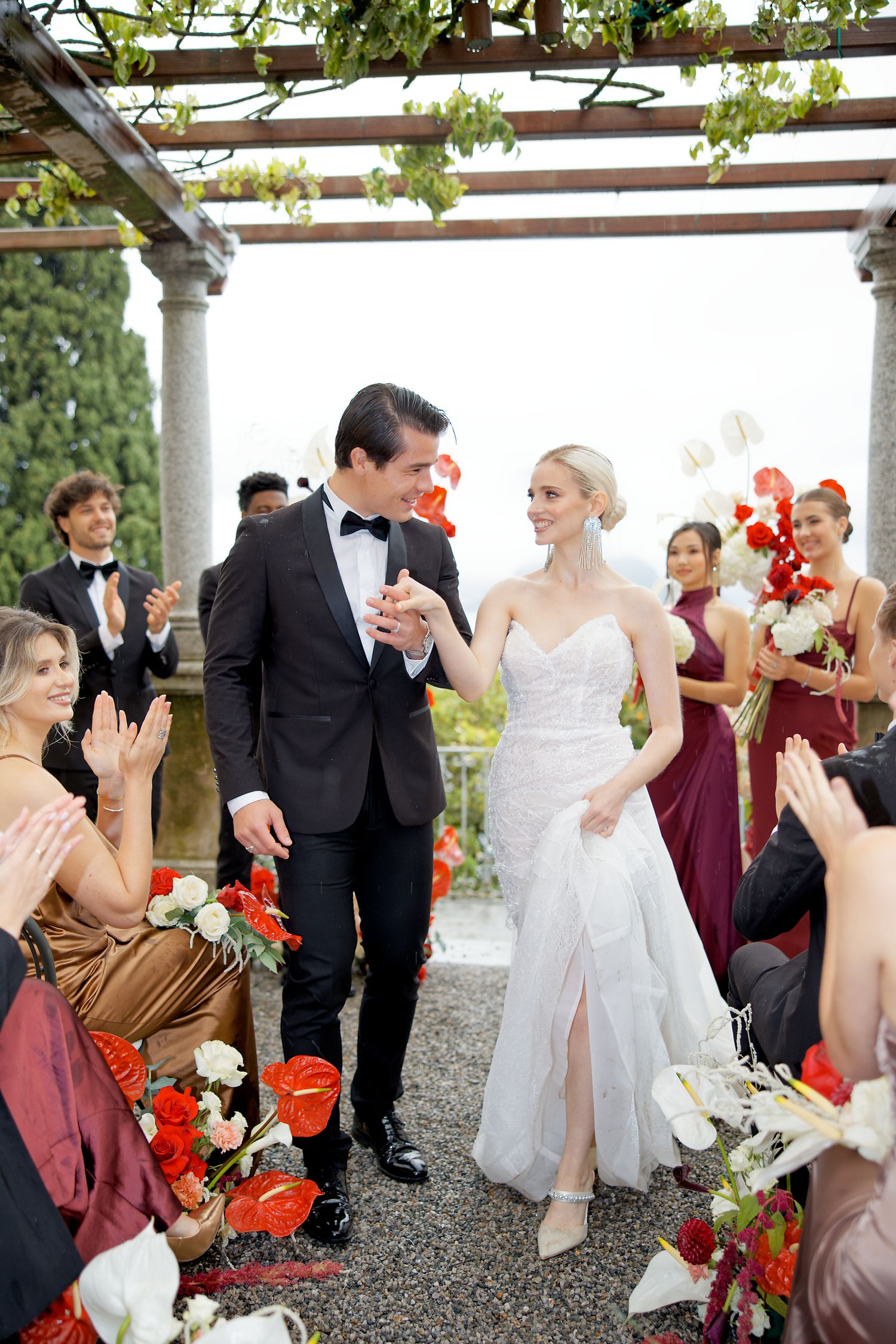 Wedding celebration under a garden pergola with guests in burgundy and gold attire.
