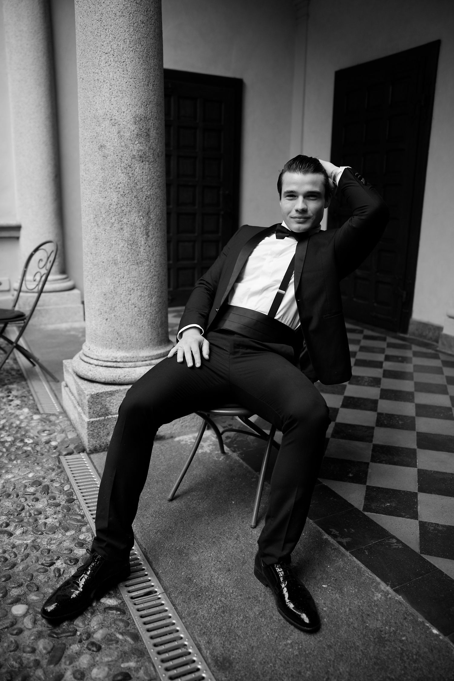 Man in tuxedo poses casually on chair against checkerboard floor and column.