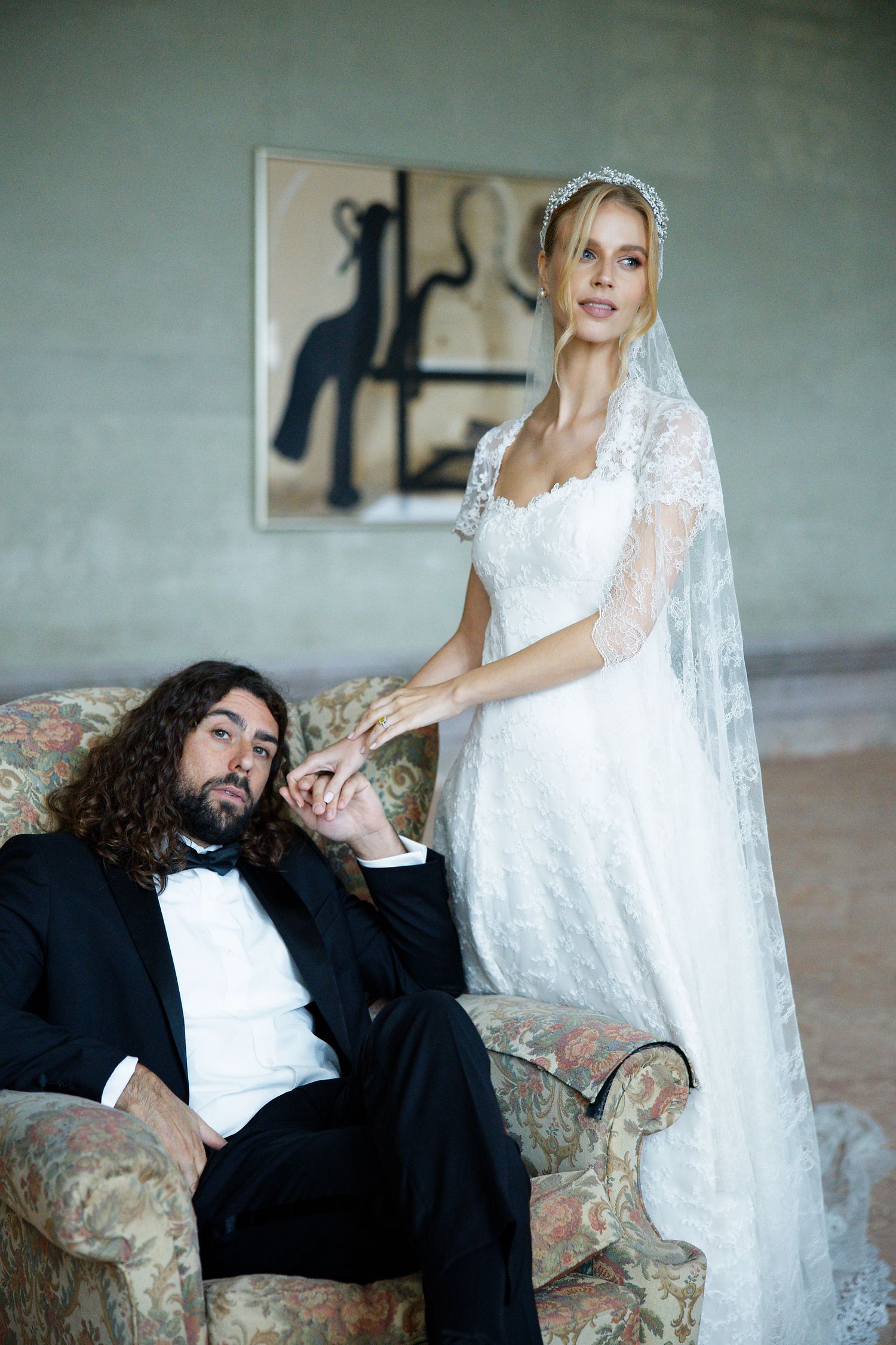 Artistic wedding photo with couple posing by antique armchair against wall with abstract artwork.