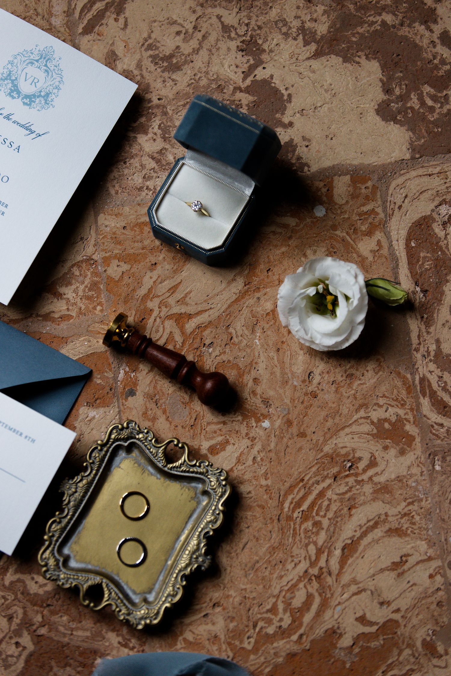 Wedding details flatlay with ring box, white flower, invitation and vintage frame on marble surface.
