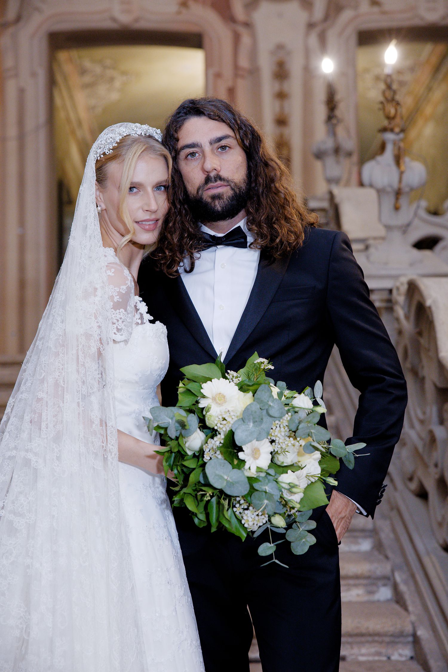 Newlywed couple with white floral bouquet standing in ornate baroque palace hallway.