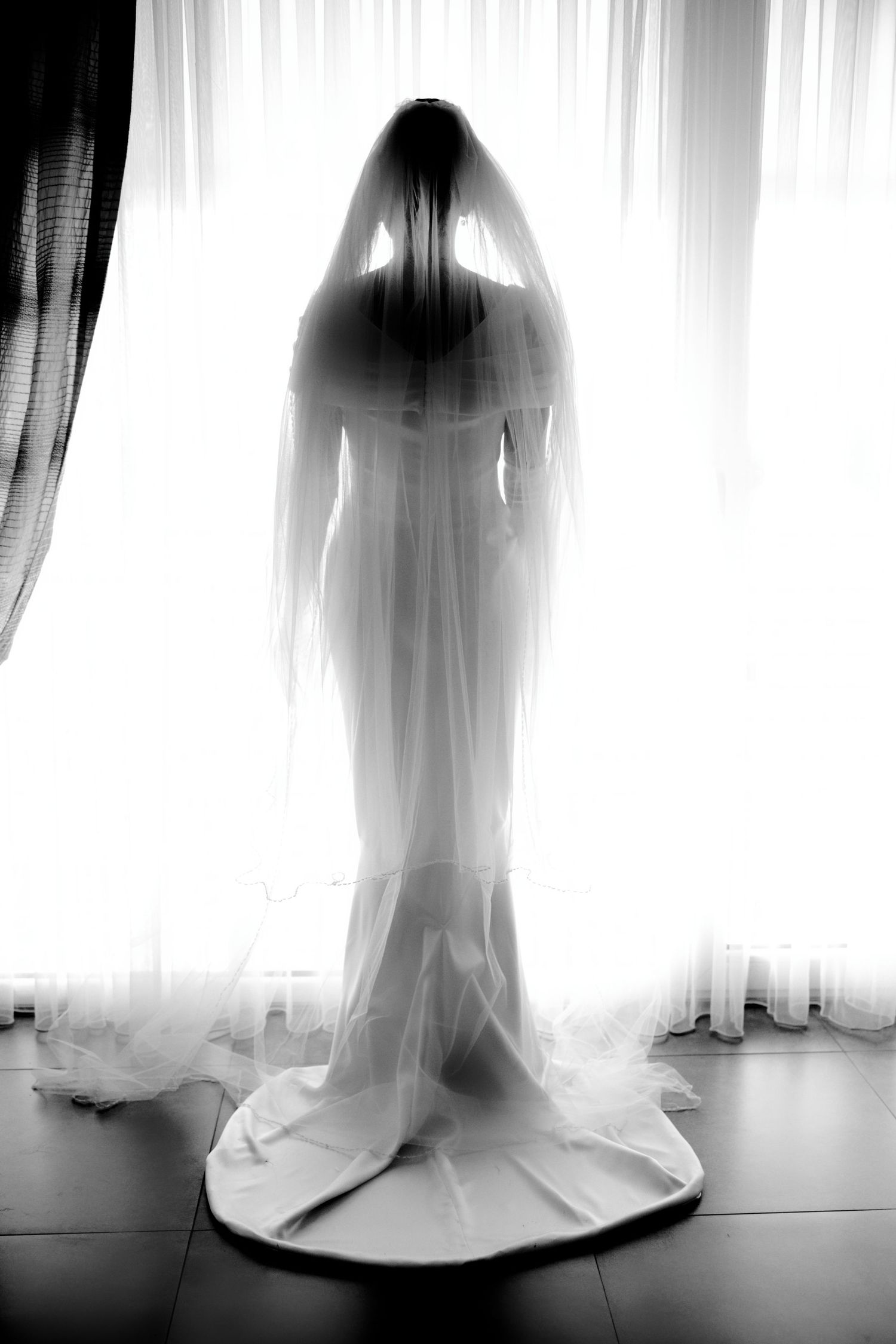 Silhouette of a person wearing a flowing wedding veil and gown standing in front of white curtains.