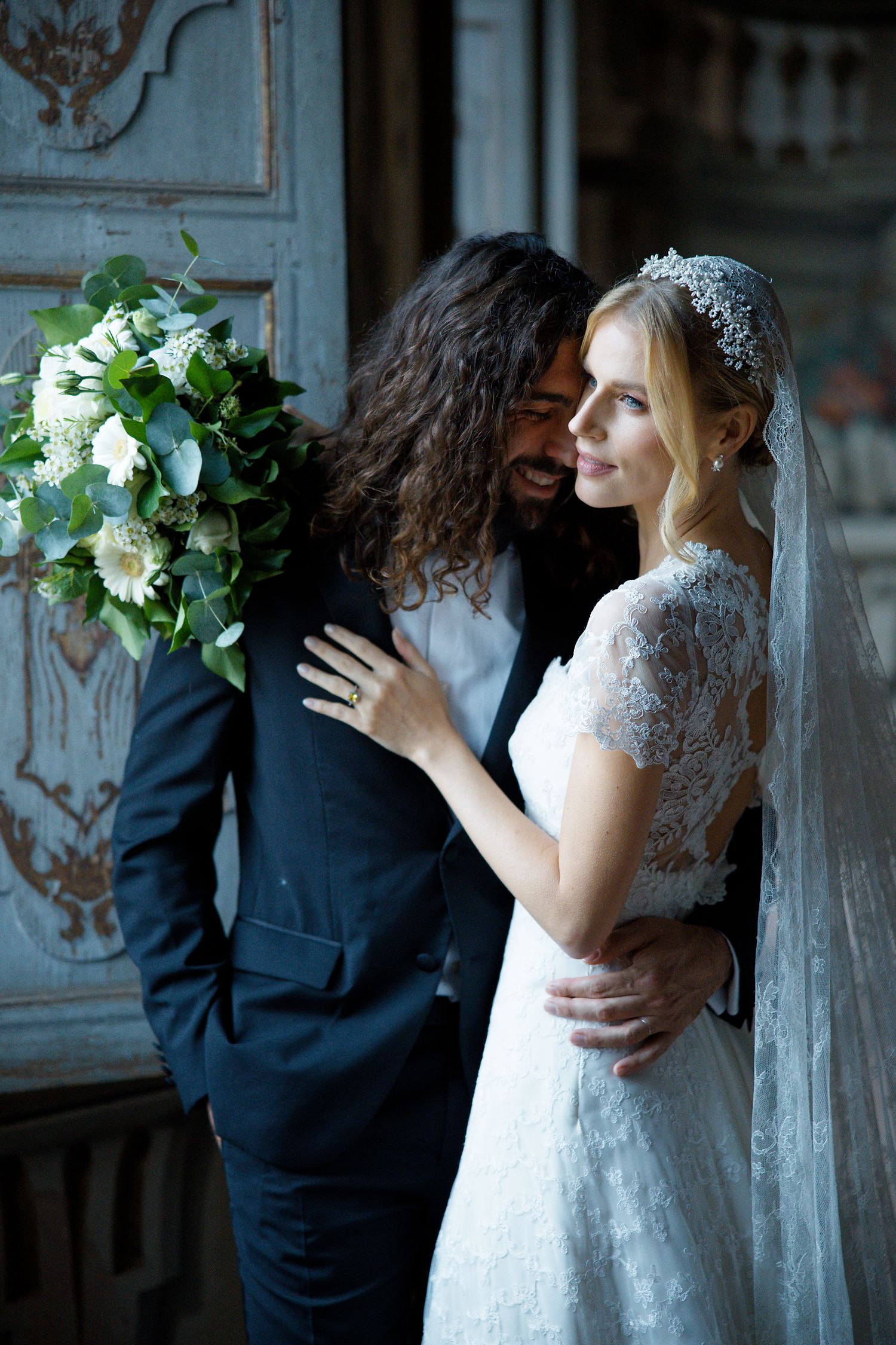 Elegant wedding couple poses by vintage blue doors with white floral bouquet.